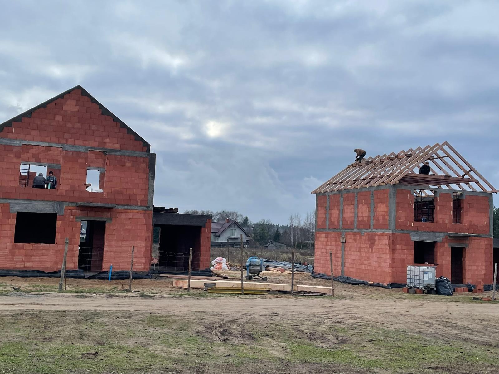Dwa domy w trakcie budowy z czerwonej cegły, jeden z ukończoną konstrukcją dachu, drugi w stanie surowym, pracownicy widoczni w oknach i na dachu, pochmurne niebo.