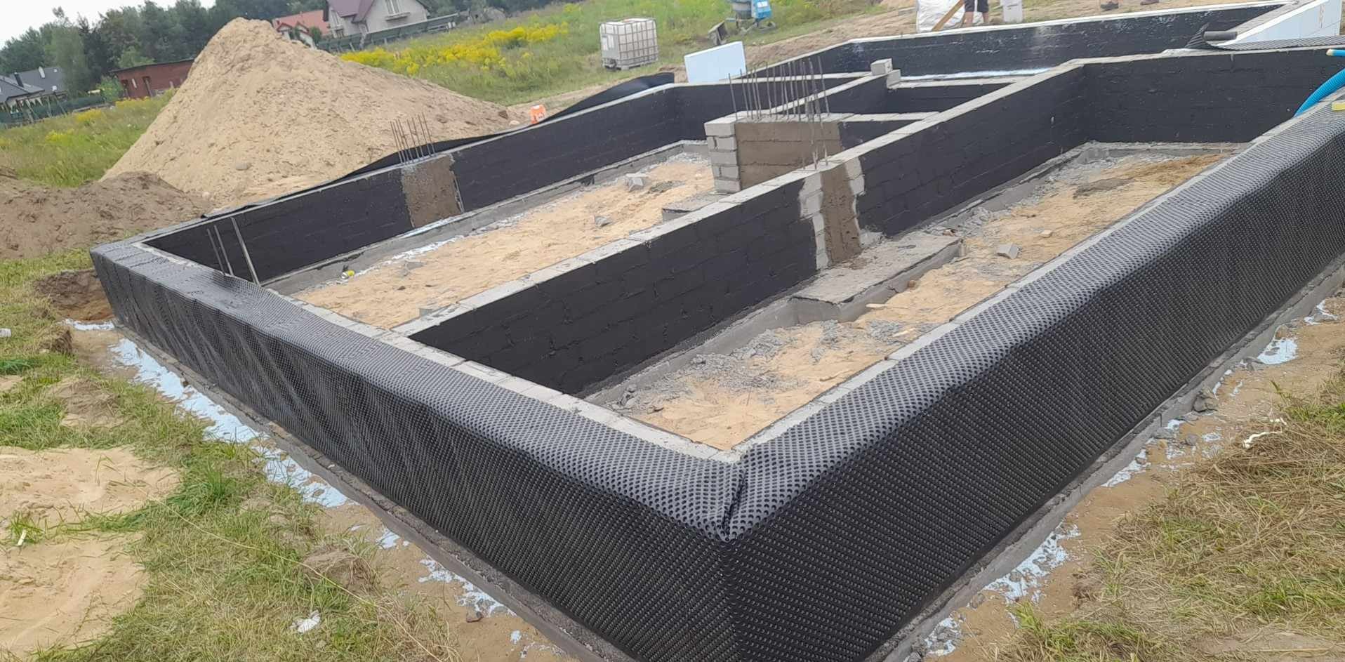 Fundamenty domu w trakcie budowy, zabezpieczone czarną folią kubełkową, widoczne zbrojenia i częściowo wylany beton, w tle hałda piasku.