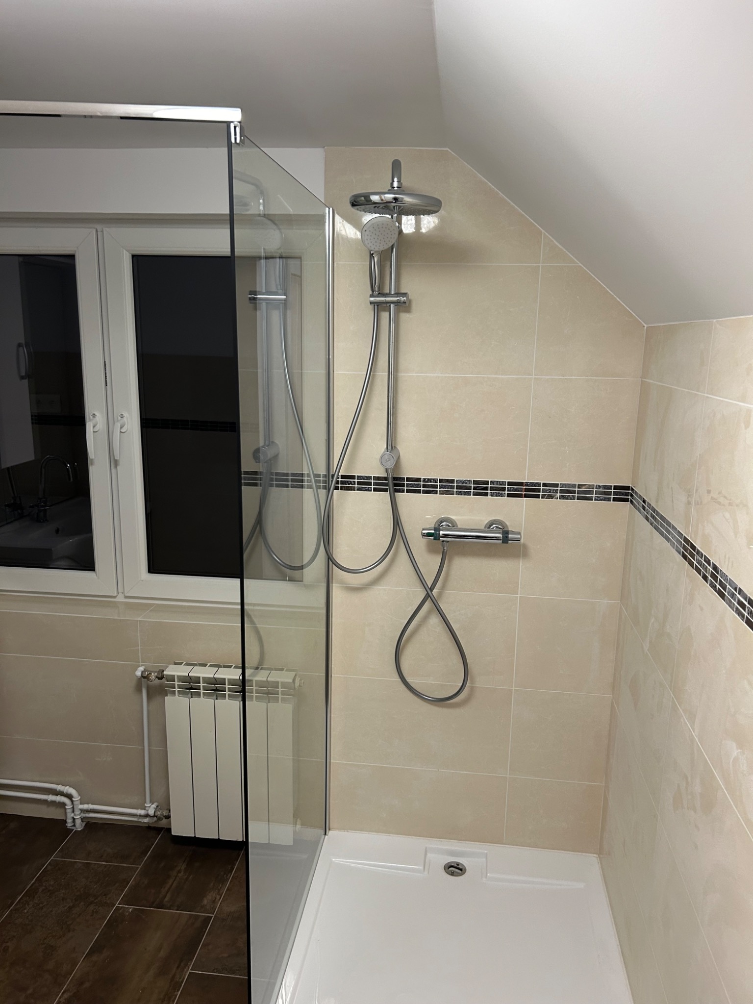 Wykończona łazienka na poddaszu z beżowymi płytkami, kabiną prysznicową ze szkła hartowanego, grzejnikiem pod oknem i czarnym paskiem dekoracyjnym na ścianach.