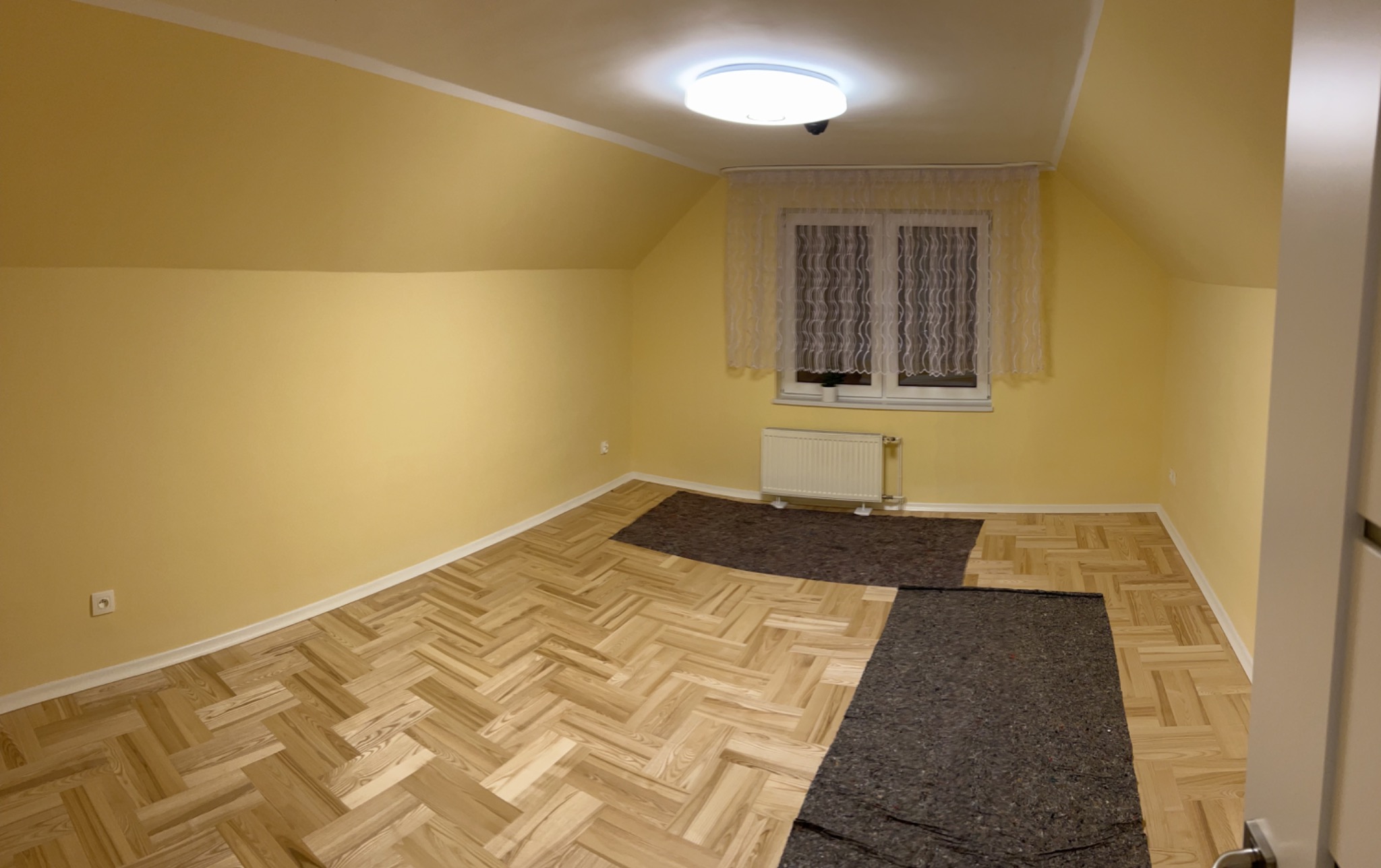 Pokój na poddaszu z żółtymi ścianami i podłogą w jodełkę, częściowo przykrytą ciemną folią ochronną, okno z firanką i grzejnikiem.