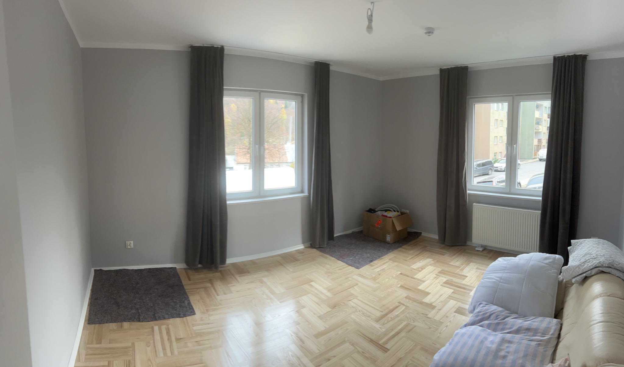 Pokój po odświeżeniu ścian w odcieniach szarości w Nowym Sączu, z parkietem w jodełkę, zasłoniętymi oknami i częściowo zakrytym meblami.