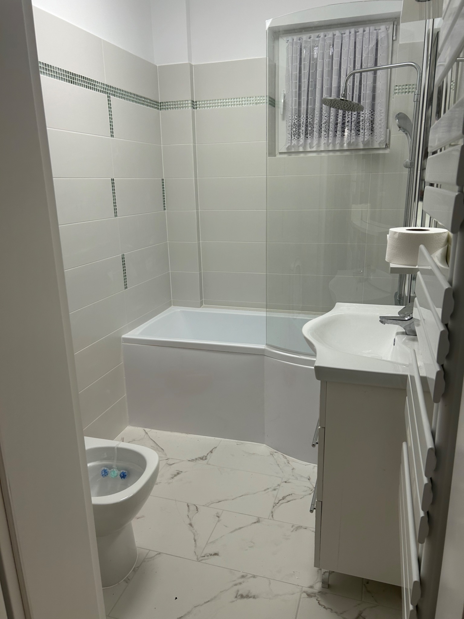 Jasna łazienka z wanną, umywalką z szafką i toaletą, wykończona białymi płytkami z dekoracyjnym paskiem mozaiki, marmurowa podłoga, grzejnik drabinkowy z papierem toaletowym.