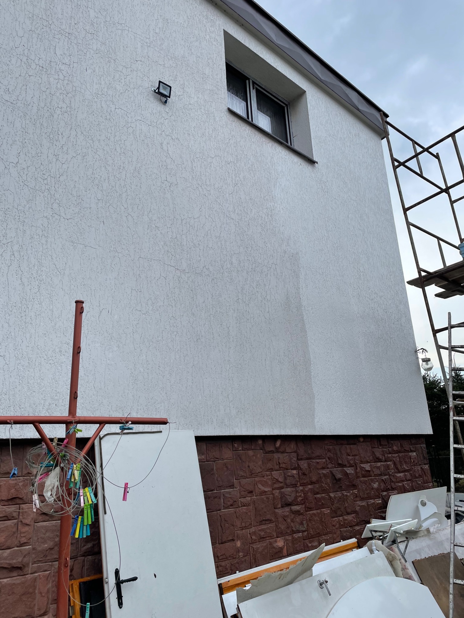 Boczna ściana domu z nowym, jasnym tynkiem strukturalnym, widoczny fragment rusztowania, okno z ciemną ramą, lampa zewnętrzna, oraz elementy odpadów budowlanych i suszarka na pranie