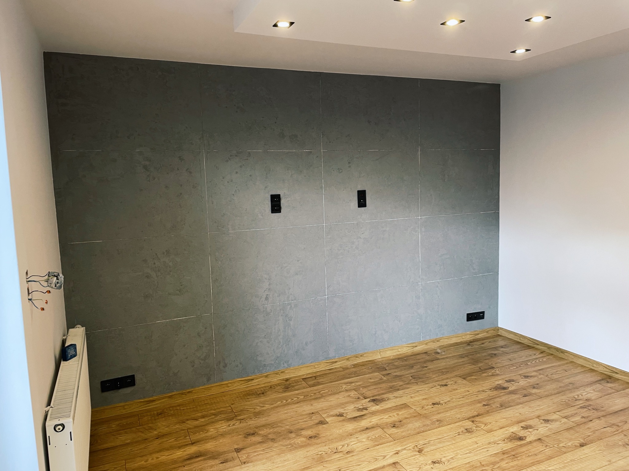 Pokój po remoncie z szarą ścianą wyłożoną płytkami imitującymi beton, drewnopodobną podłogą, białym sufitem z oświetleniem punktowym i grzejnikiem z podłączonymi przewodami elektrycznymi.
