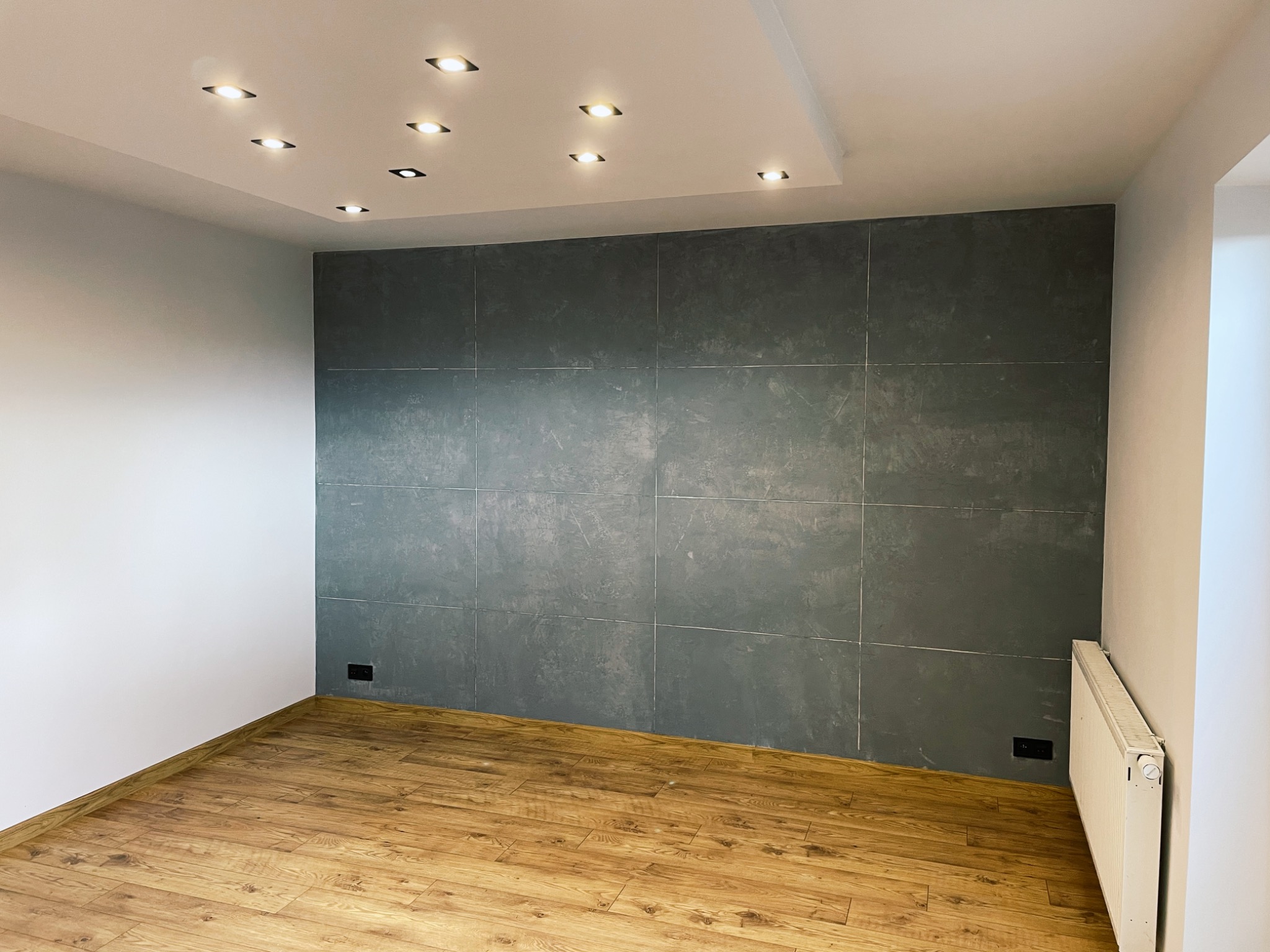 Wykończone wnętrze pokoju z drewnianą podłogą, szarą ścianą z płyt, białymi ścianami i sufitem podwieszanym z oświetleniem punktowym.