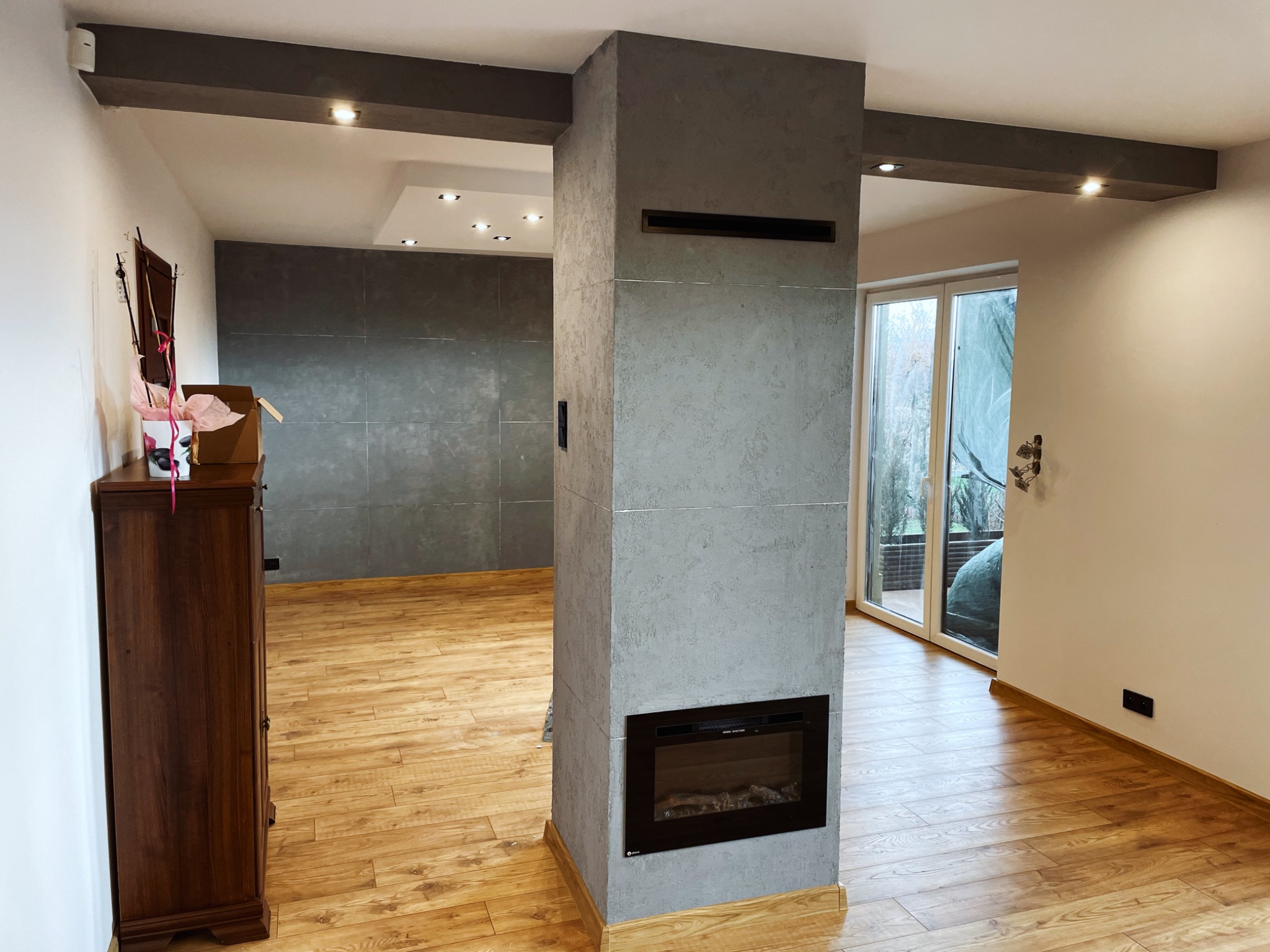 Wnętrze po remoncie z drewnianą podłogą, szarą ścianą z płyt gipsowych i wbudowanym elektrycznym kominkiem w szarym słupie, widoczne również szare belki sufitowe z oświetleniem punktowym.
