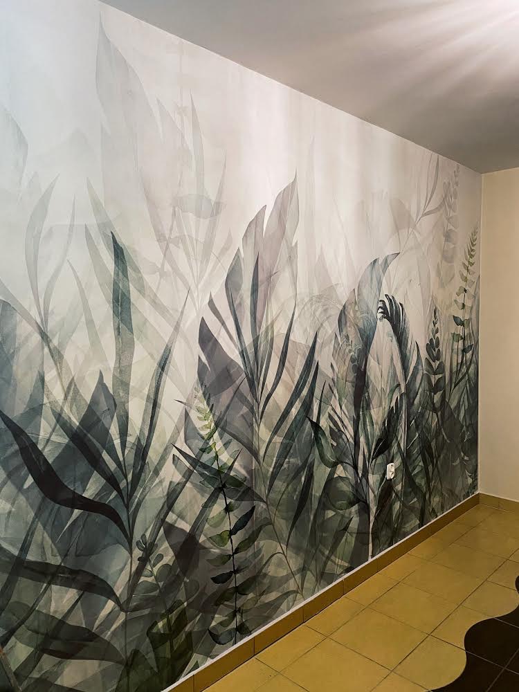Ściana w pokoju pokryta fototapetą z motywem roślin tropikalnych w odcieniach szarości i zieleni; widoczny fragment podłogi z żółtymi płytkami ceramicznymi.