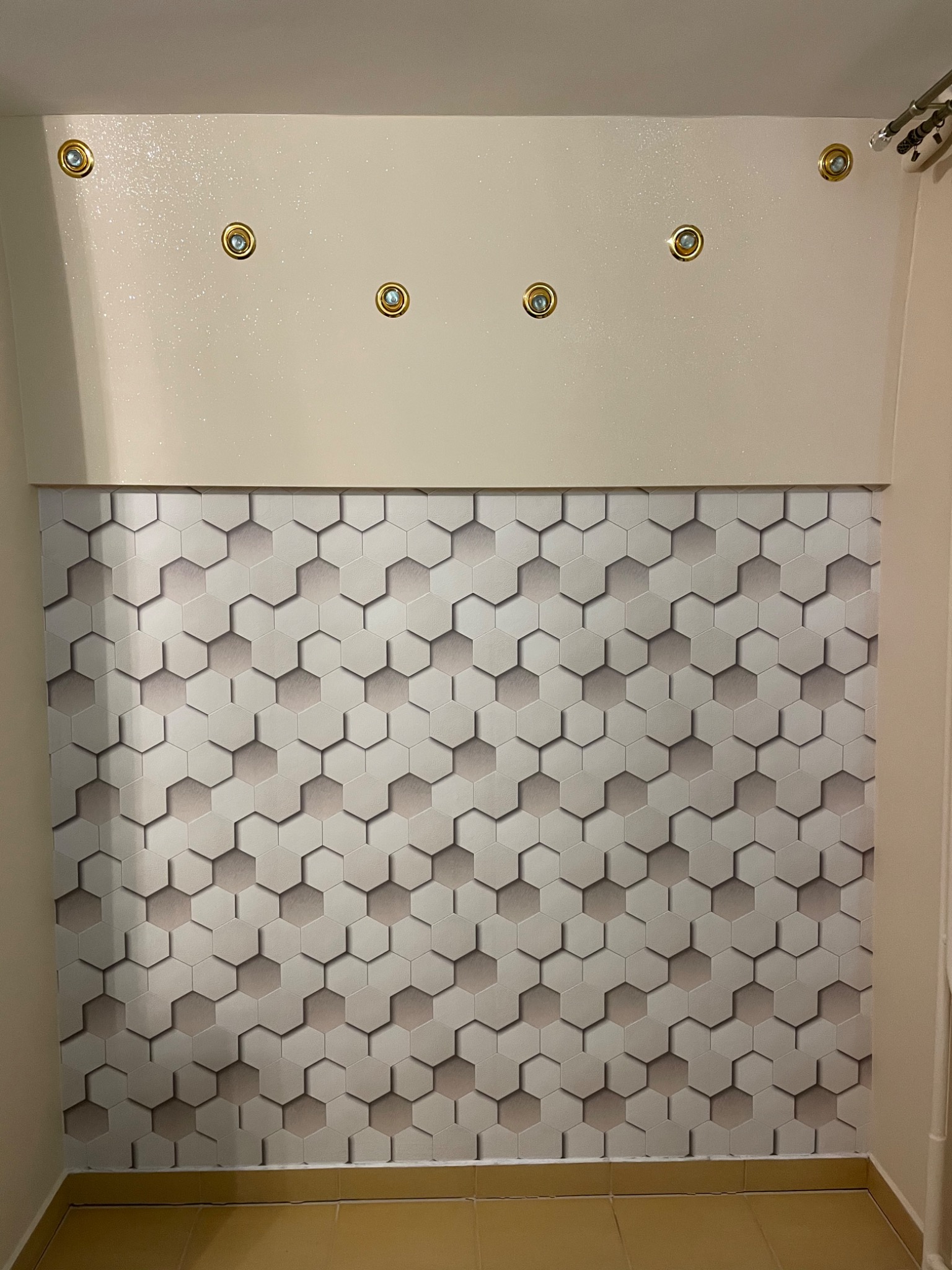Ściana z geometryczną tapetą w odcieniach szarości i beżu, oświetlona punktowymi lampami z złotymi obwódkami, zamontowanymi w gładkim, beżowym panelu sufitowym.