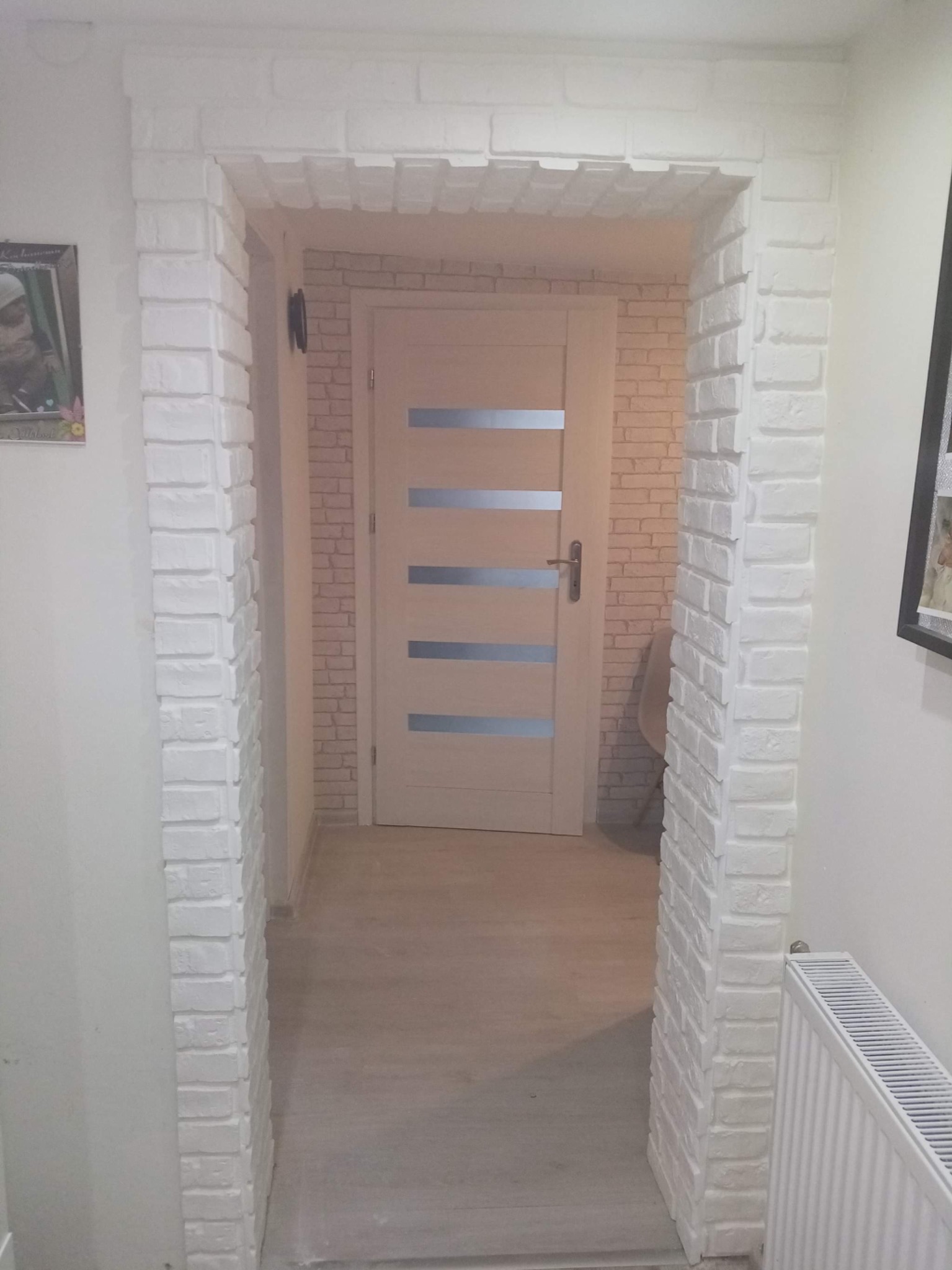 Biały, ceglany portal drzwiowy z widoczną ścianą w cegiełki i jasnymi drzwiami z poziomymi przeszkleniami, widoczne elementy wystroju wnętrza.