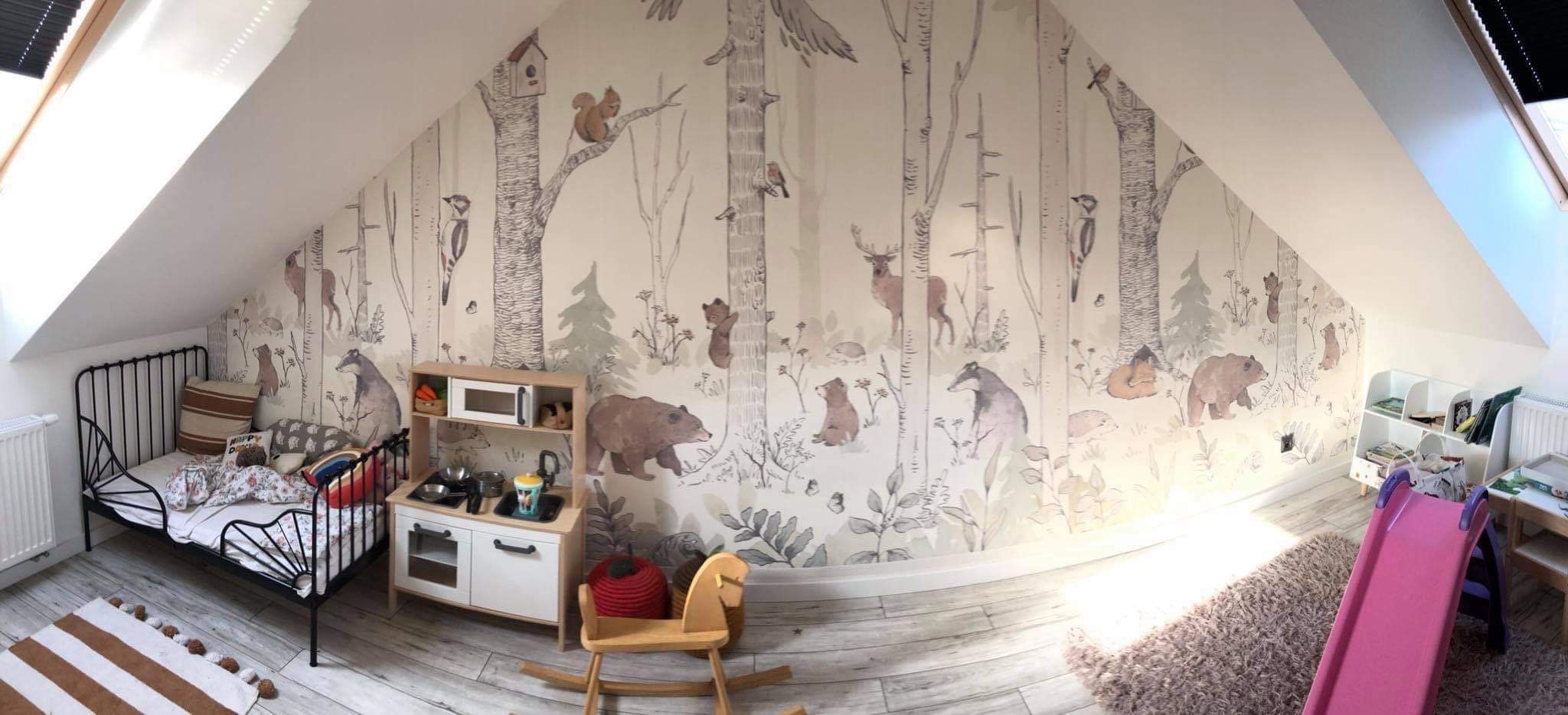 Pokój dziecięcy na poddaszu z łóżkiem, drewnianą zabawką konikiem, regałem i ścianą pokrytą tapetą z motywem leśnym, przedstawiającą drzewa, zwierzęta takie jak niedźwiedzie, wiewiórki, sarny i ptaki.