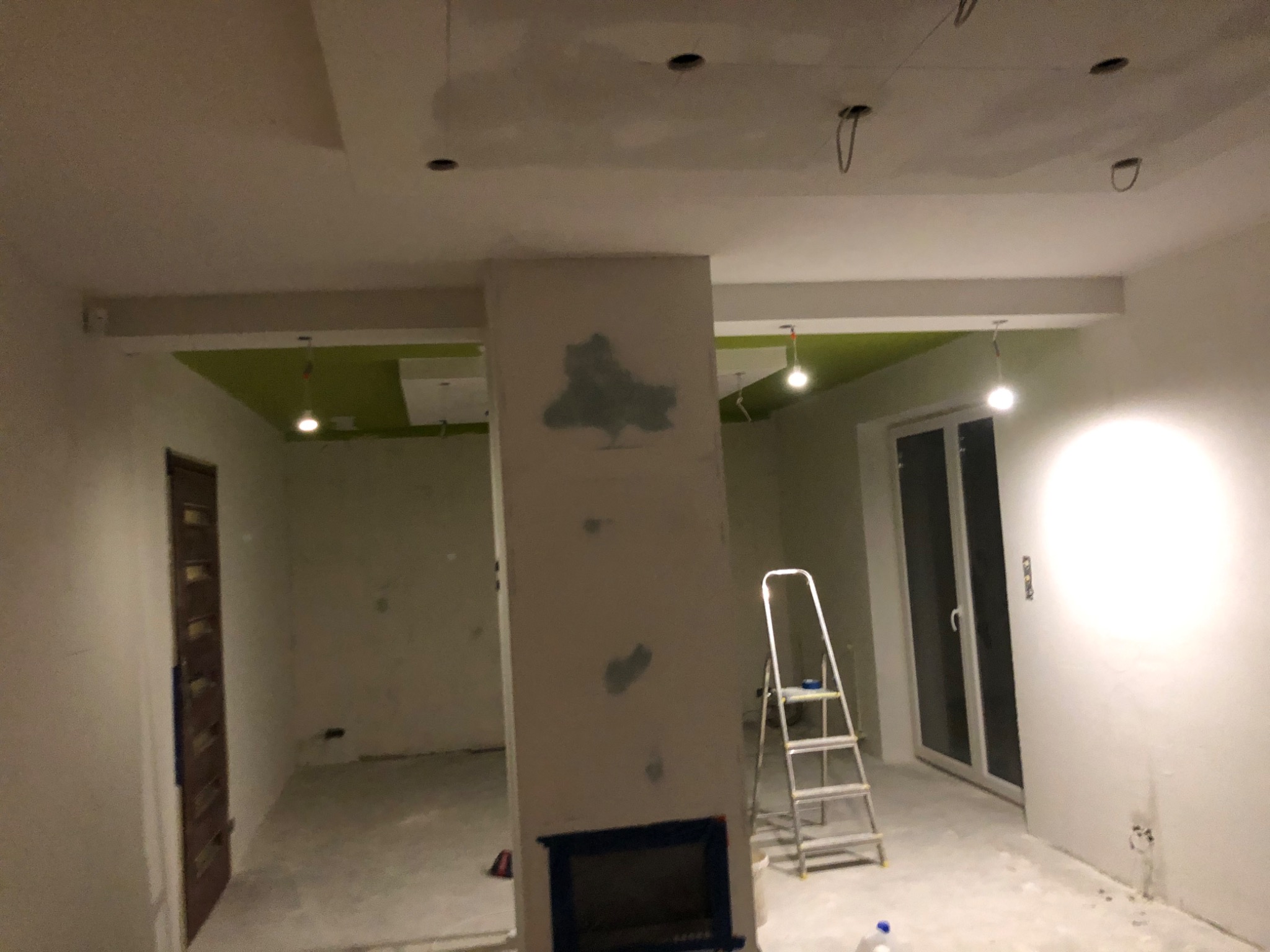 Wnętrze w trakcie remontu: sufit z punktowymi lampami, ściany przygotowane pod gładzie, widoczny kominek z zabezpieczonym otworem, drabina malarska i zielony fragment sufitu podwieszanego.