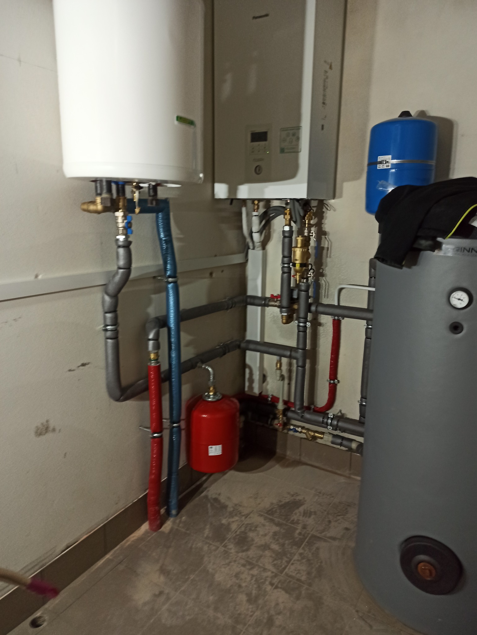 Złożona instalacja grzewcza z kotłem dwufunkcyjnym, zasobnikiem wody i naczyniami przeponowymi, rury w izolacji, widoczne liczne zawory i przyłącza.