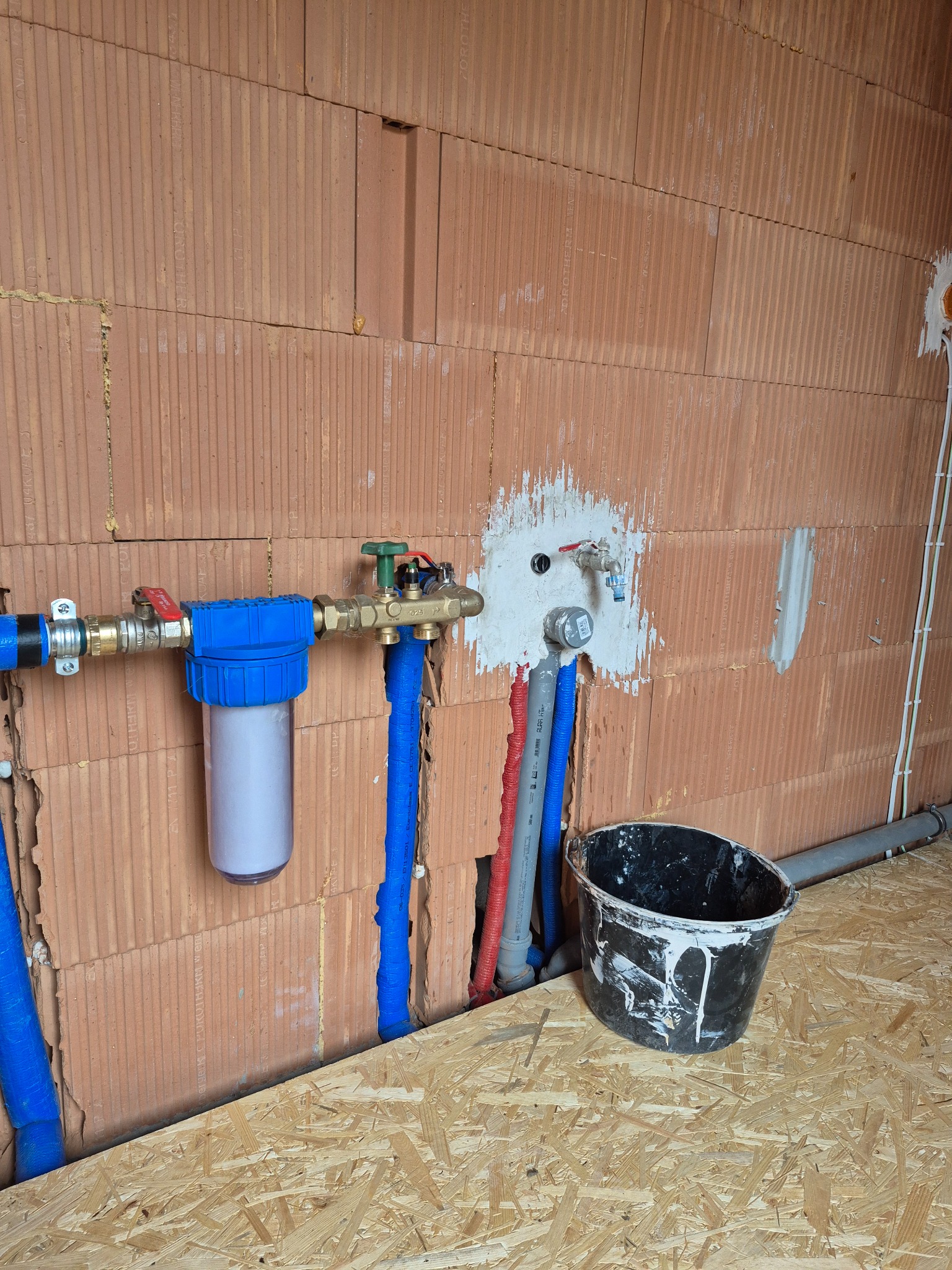 Instalacja wodna z filtrem i rurami w ścianie z czerwonej cegły. Widoczny fragment podłogi z płyty OSB i wiadro. Prace hydrauliczne w trakcie realizacji.