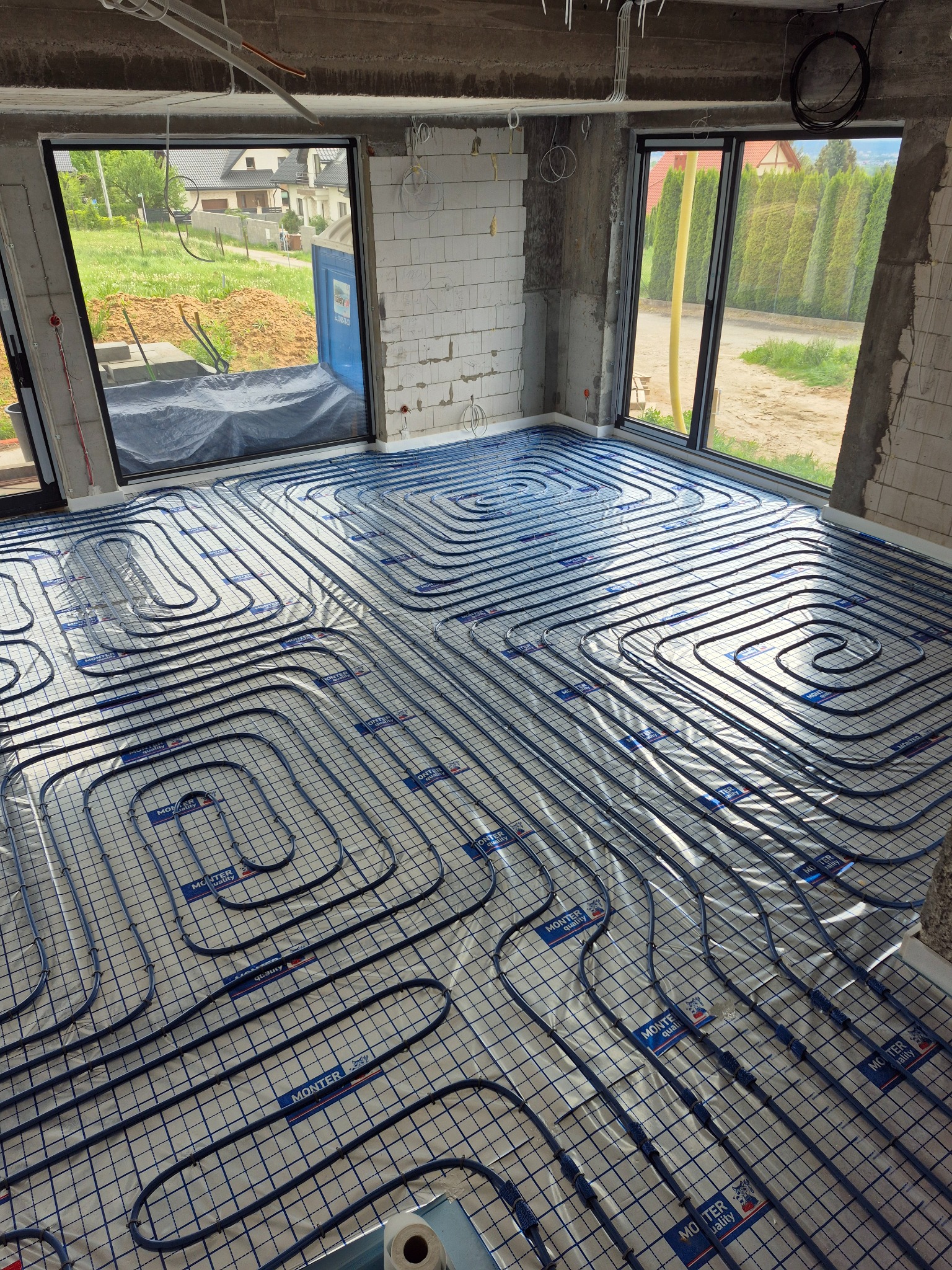 Rozłożona instalacja ogrzewania podłogowego w surowym wnętrzu z dużymi oknami. Widoczne rury w charakterystycznym układzie na folii izolacyjnej.