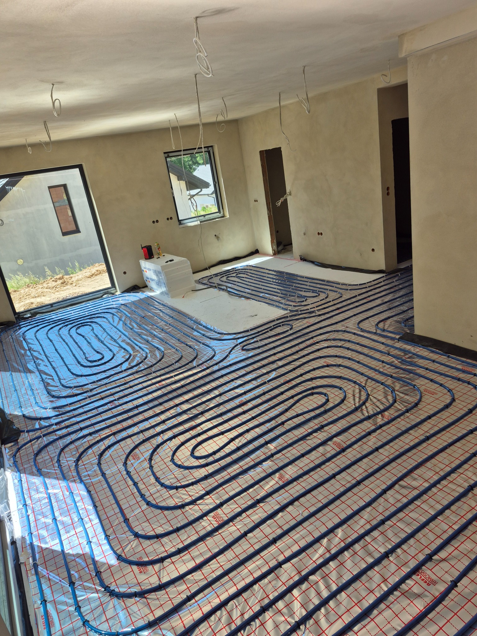 Niebieskie rury ogrzewania podłogowego na folii termoizolacyjnej w surowym wnętrzu z oknami i instalacją elektryczną na suficie. Widok z góry na rozłożoną instalację.