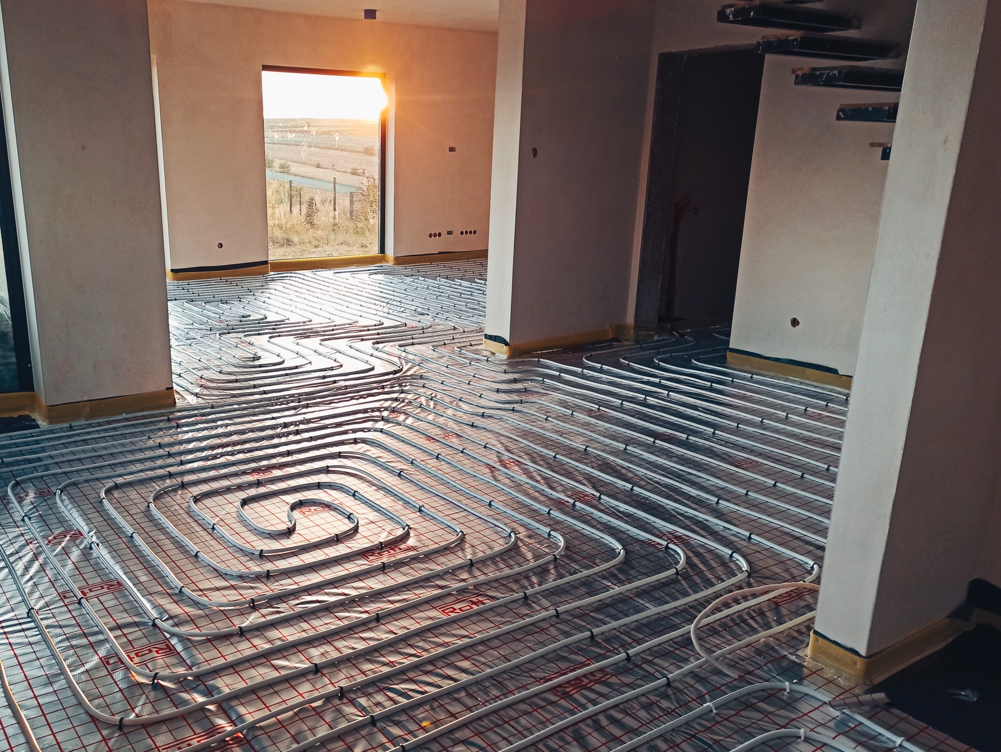 Białe rury ogrzewania podłogowego ułożone spiralnie na folii z czerwoną siatką w jasnym, surowym wnętrzu domu w trakcie budowy, z widokiem na zewnątrz przez okno.