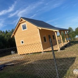 MARDOM-DOMY DREWNIANE - Nowo wybudowany, jasny, drewniany domek letniskowy z ciemnym dachem, widoczny zza ogrodzenia z siatki, z elementami wykończenia tarasu.