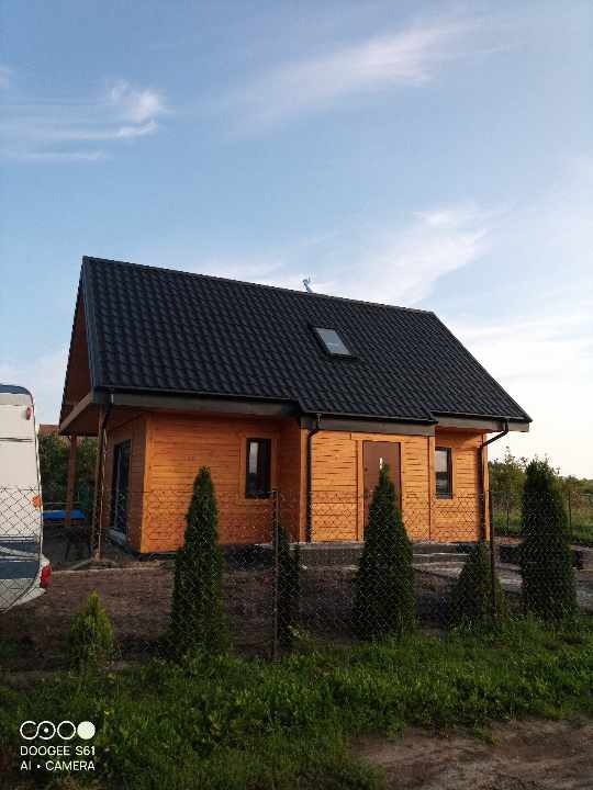 Gotowy, drewniany dom z czarnym dachem, widoczny z zewnątrz, otoczony młodymi tujami i ogrodzeniem z siatki, z przyczepą kempingową w tle.