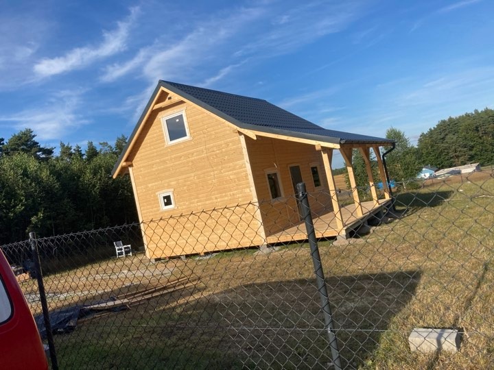 Nowo wybudowany, jasny, drewniany domek letniskowy z ciemnym dachem, widoczny zza ogrodzenia z siatki, z elementami wykończenia tarasu.