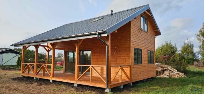 Gotowy, parterowy dom z drewna z zadaszonym tarasem, widoczne czarne rynny i okna, w tle sterta drewna i zieleń.