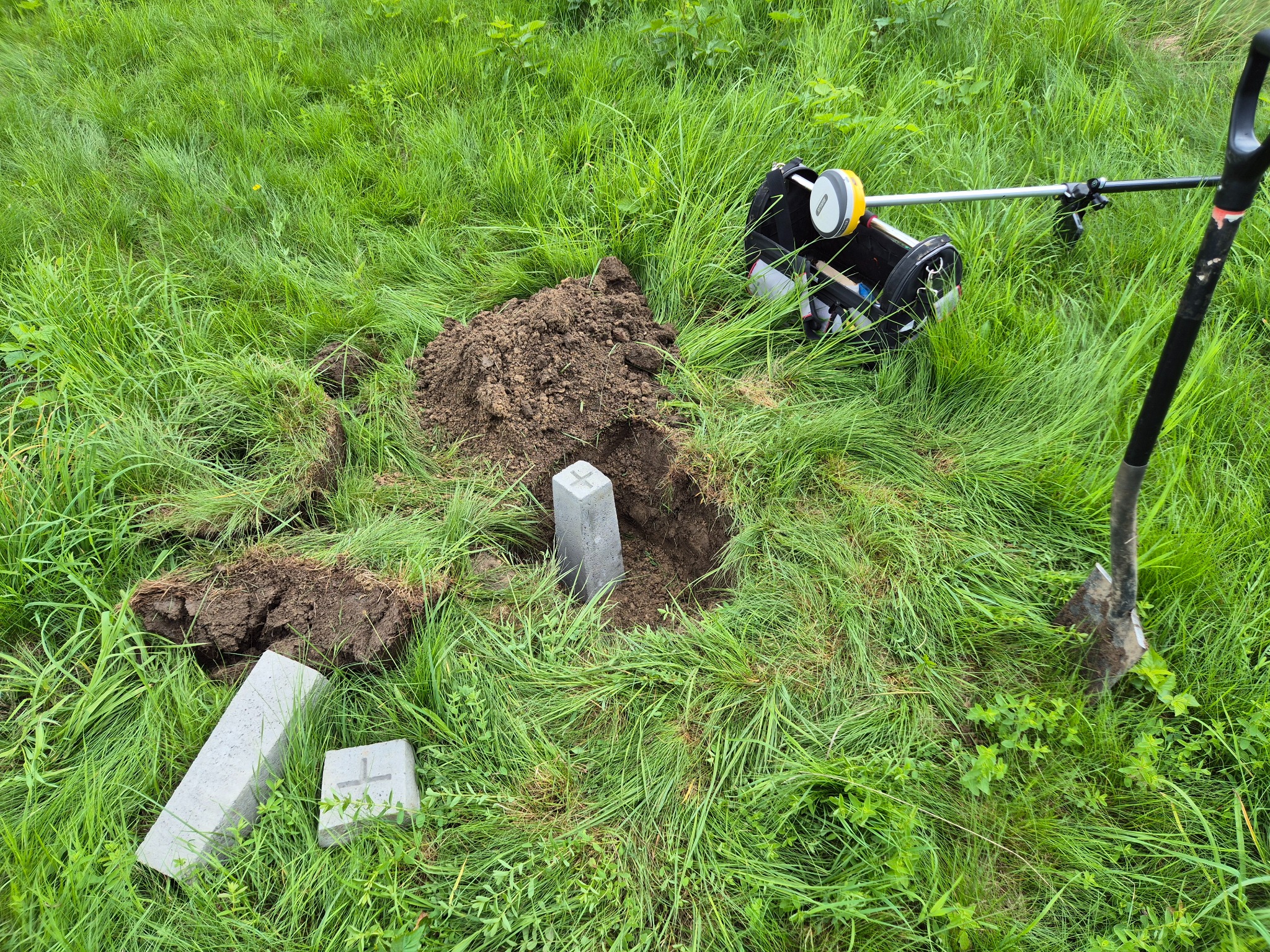 Prace geodezyjne w terenie: wykopany dół, betonowy słupek graniczny z krzyżem, sprzęt pomiarowy i łopata na zarośniętej trawie. Stabilizacja punktów granicznych.