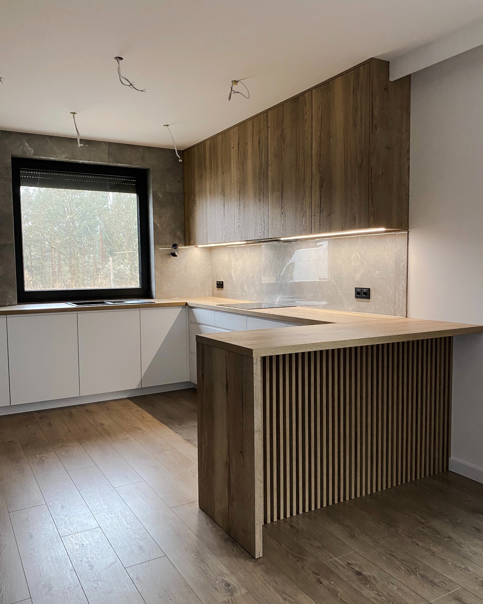 Nowoczesna kuchnia z białymi szafkami dolnymi i drewnopodobnymi górnymi, wyspa z lamelami, okno z czarną ramą, widoczne niedokończone podłączenia elektryczne na suficie.