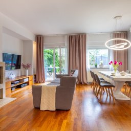 DOMUS NIERUCHOMOŚCI HANNA MORAWSKA - Jasne, przestronne wnętrze salonu z drewnianą podłogą, beżowym kominkiem, szarą sofą z narzutą i białym stołem jadalnym z kwiatami.