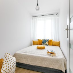 DOMUS NIERUCHOMOŚCI HANNA MORAWSKA - Mała, przytulna sypialnia z niskim łóżkiem, żółtymi poduszkami, książkami i okularami na beżowej narzucie, widok na białą ścianę i okno z białymi zasłonami, kosz z poduszkami w rogu.