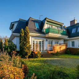 DOMUS NIERUCHOMOŚCI HANNA MORAWSKA - Dwupiętrowy dom z szarego kamienia z ciemnym dachem, balkonem z zieloną balustradą i zadbanym ogrodem z drzewami iglastymi i krzewami.