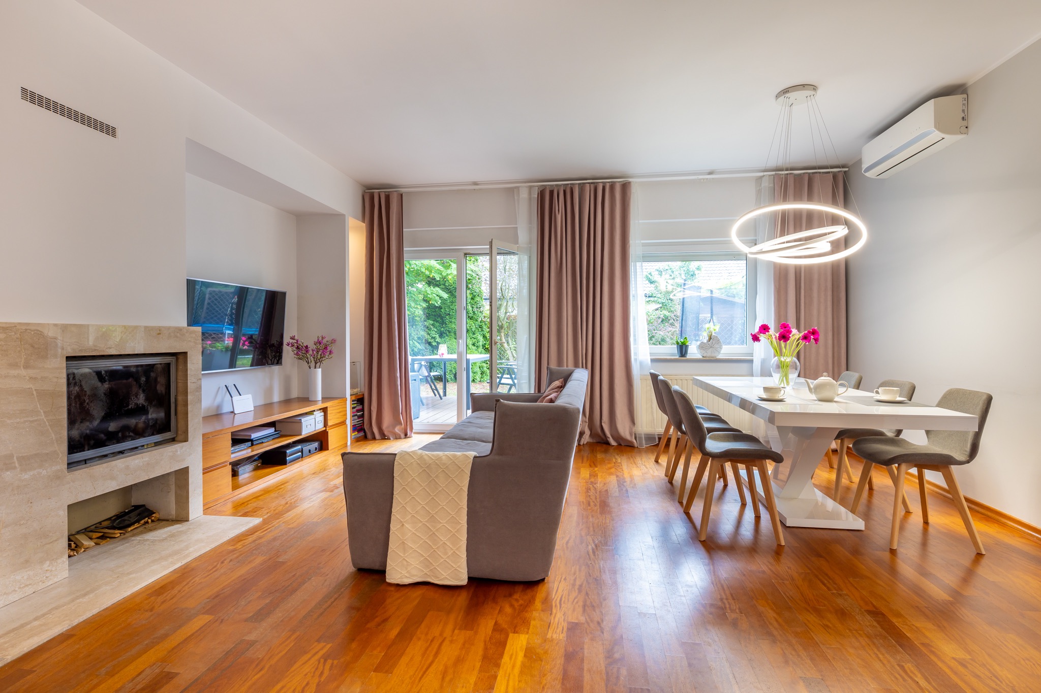 Jasne, przestronne wnętrze salonu z drewnianą podłogą, beżowym kominkiem, szarą sofą z narzutą i białym stołem jadalnym z kwiatami.