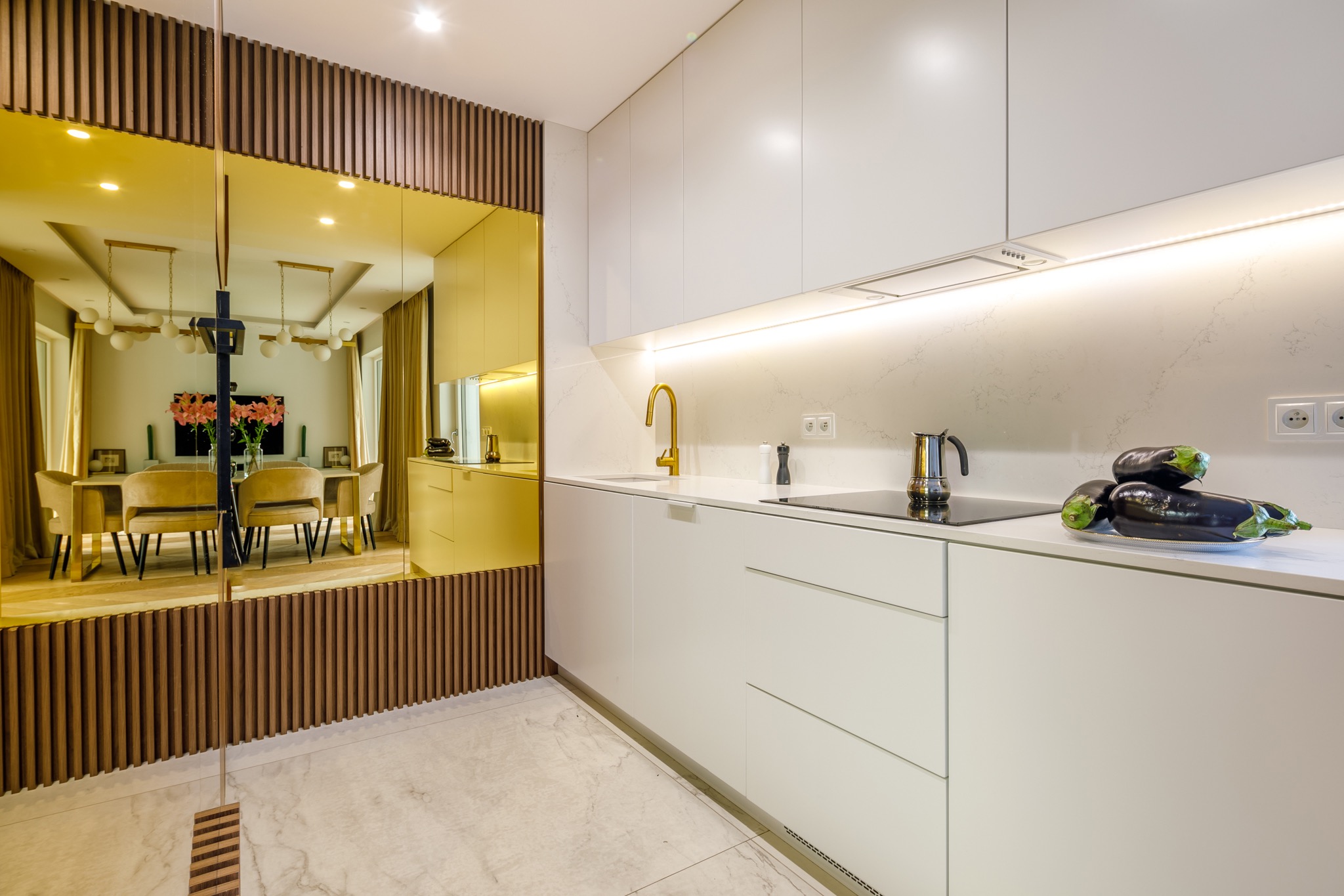 Nowoczesna, minimalistyczna kuchnia z białymi szafkami, złotą armaturą i lustrzaną ścianą z drewnianymi listwami, w której odbija się jadalnia z kwiatami na stole.