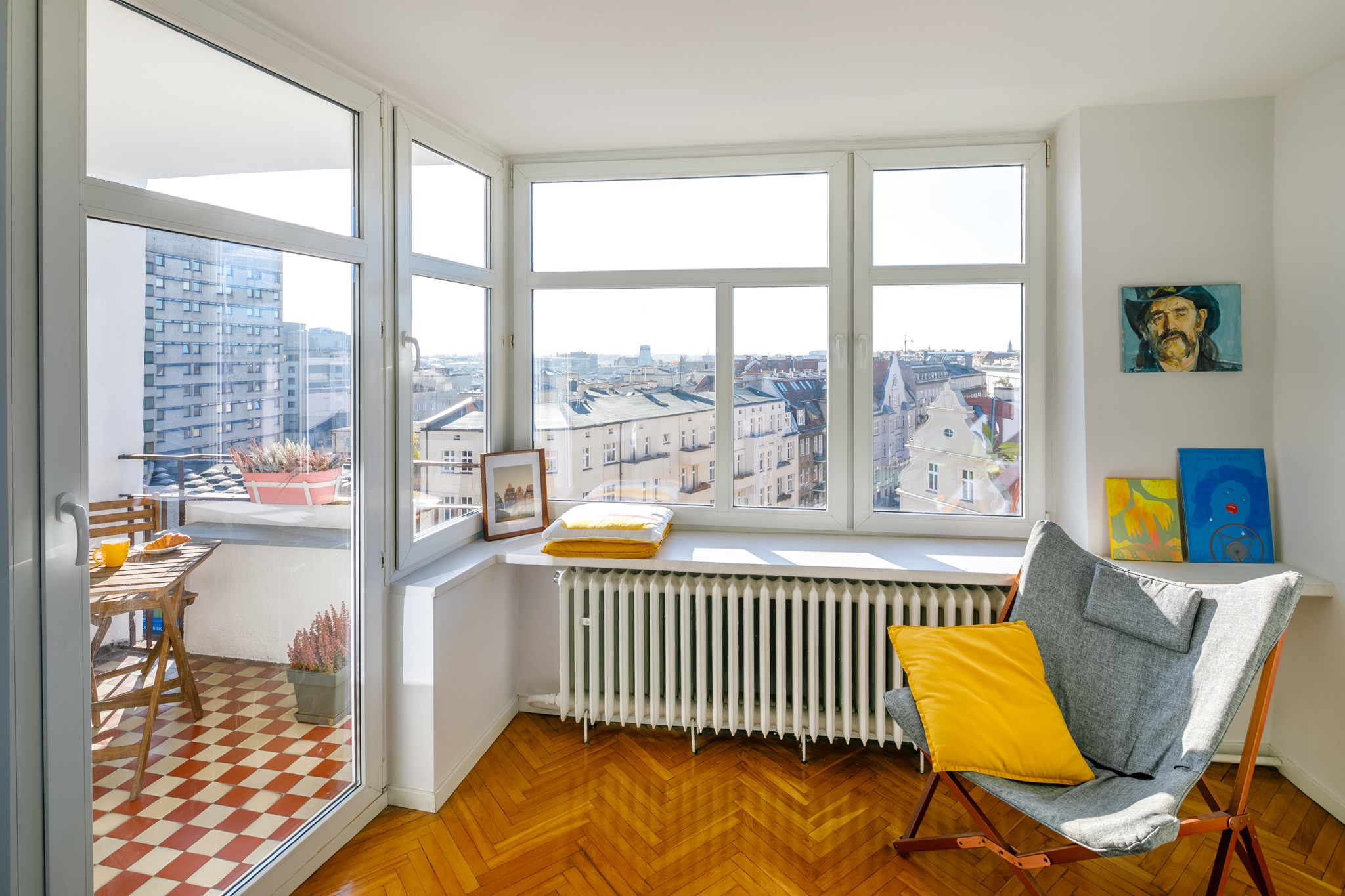 Jasne wnętrze mieszkania z widokiem na miasto z balkonu, drewniana podłoga, szare krzesło z żółtą poduszką, obrazy na ścianie, stół z jedzeniem na balkonie.