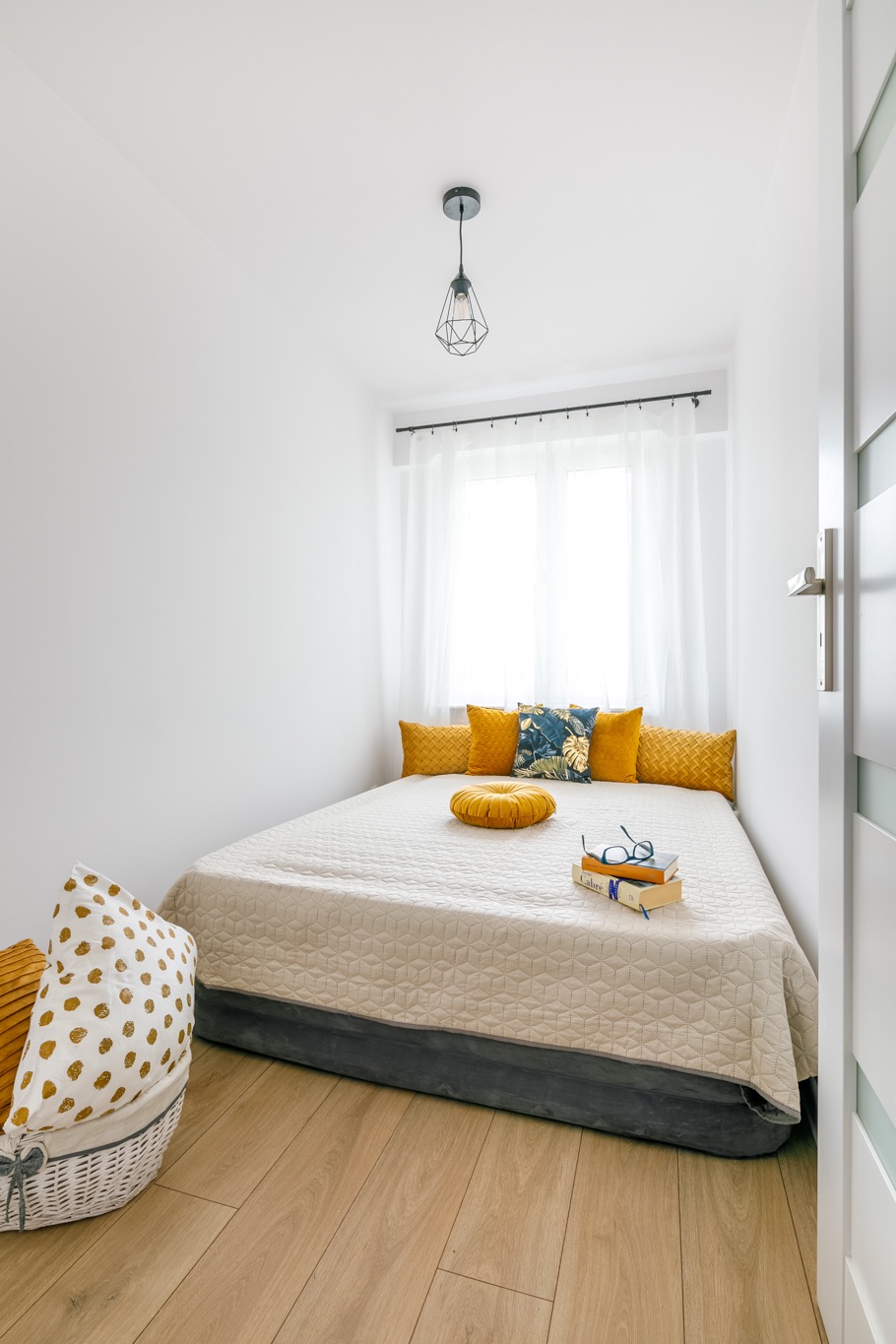 Mała, przytulna sypialnia z niskim łóżkiem, żółtymi poduszkami, książkami i okularami na beżowej narzucie, widok na białą ścianę i okno z białymi zasłonami, kosz z poduszkami w rogu.