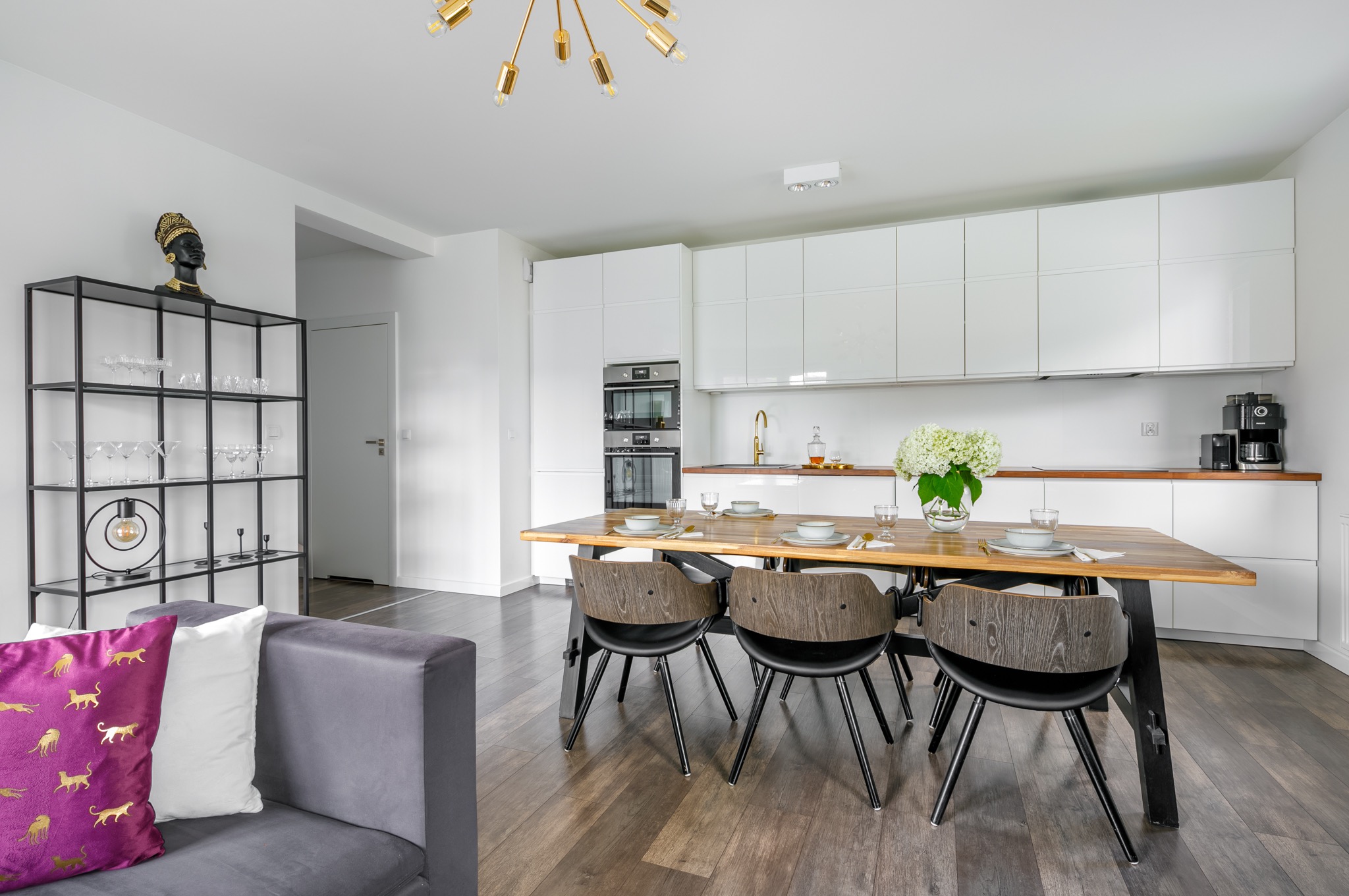 Jasne, nowoczesne wnętrze: stół z nakryciem, białe szafki kuchenne, regał z dekoracjami i szara sofa z fioletową poduszką w złote pantery.
