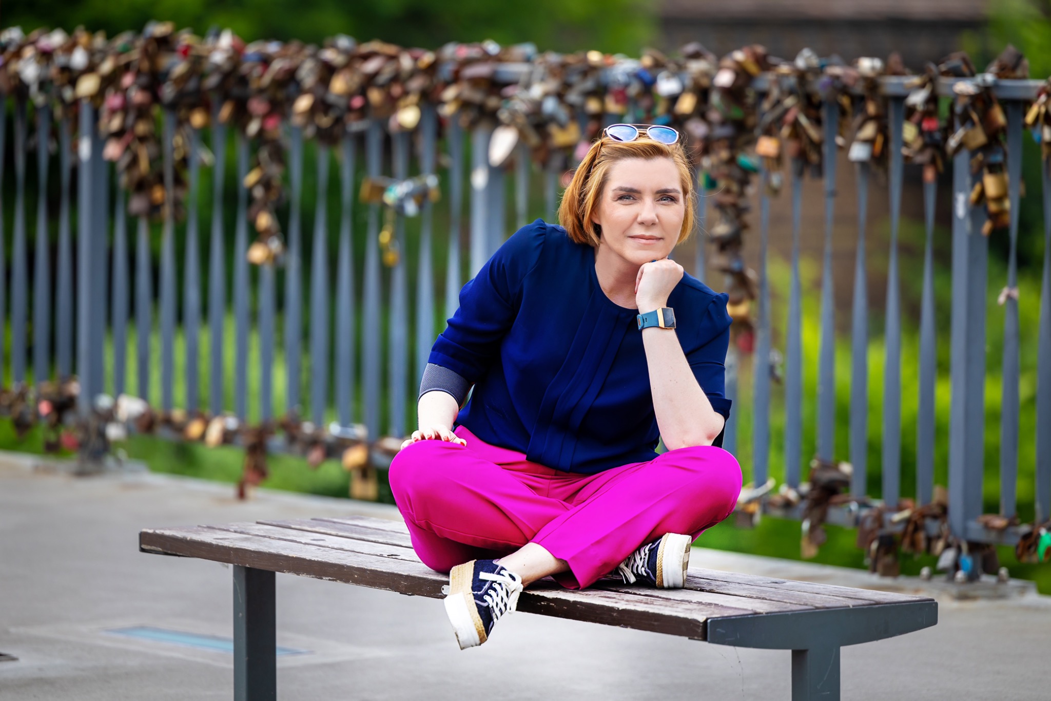 Kobieta z rudymi włosami w okularach na głowie, ubrana w granatową bluzkę i różowe spodnie, siedzi ze skrzyżowanymi nogami na ławce przed mostem ozdobionym kłódkami.