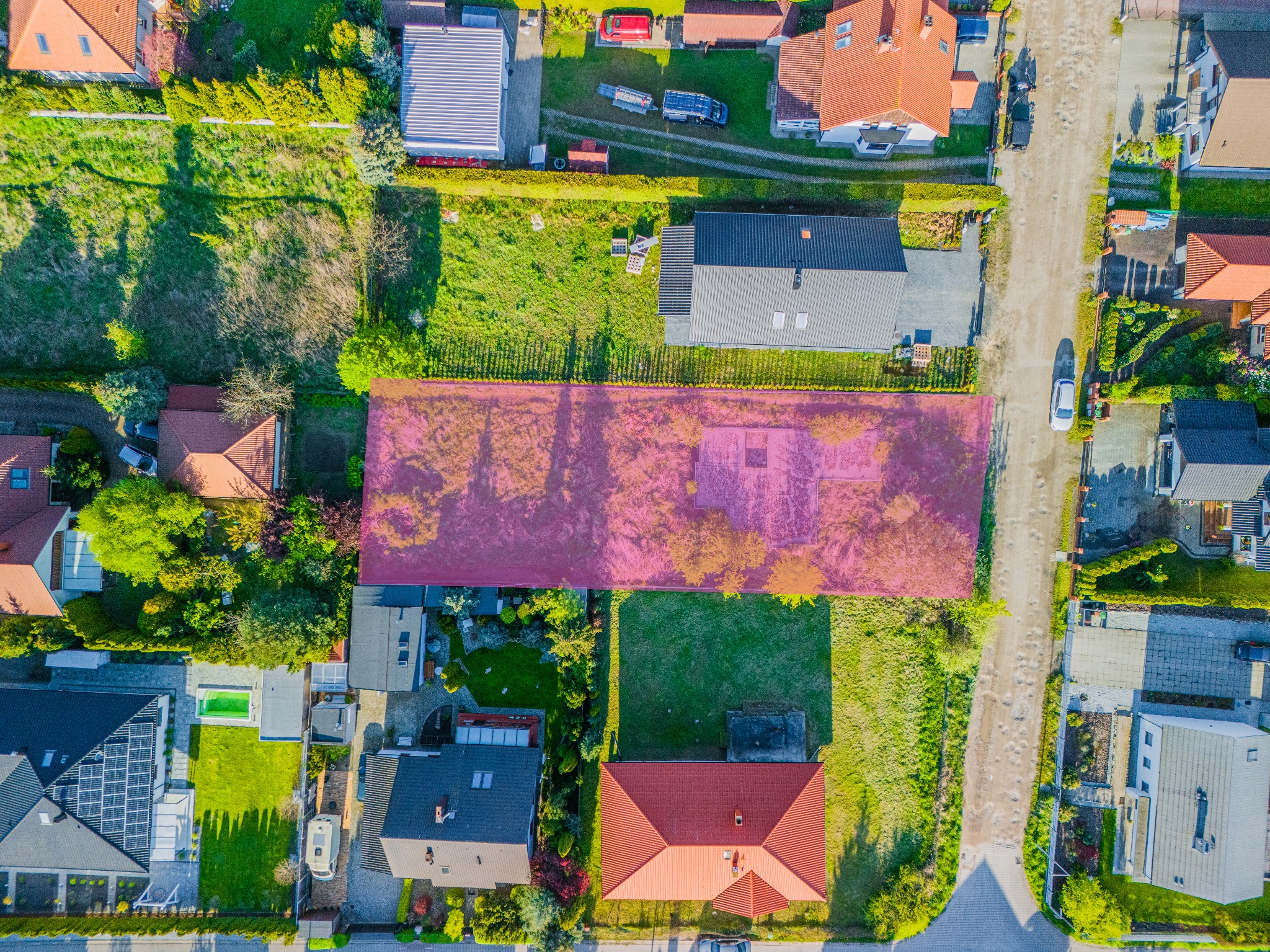 Zdjęcie z lotu ptaka przedstawia działkę budowlaną otoczoną zabudową jednorodzinną, zaznaczoną różową obwódką, z widoczną drogą dojazdową i zielenią.