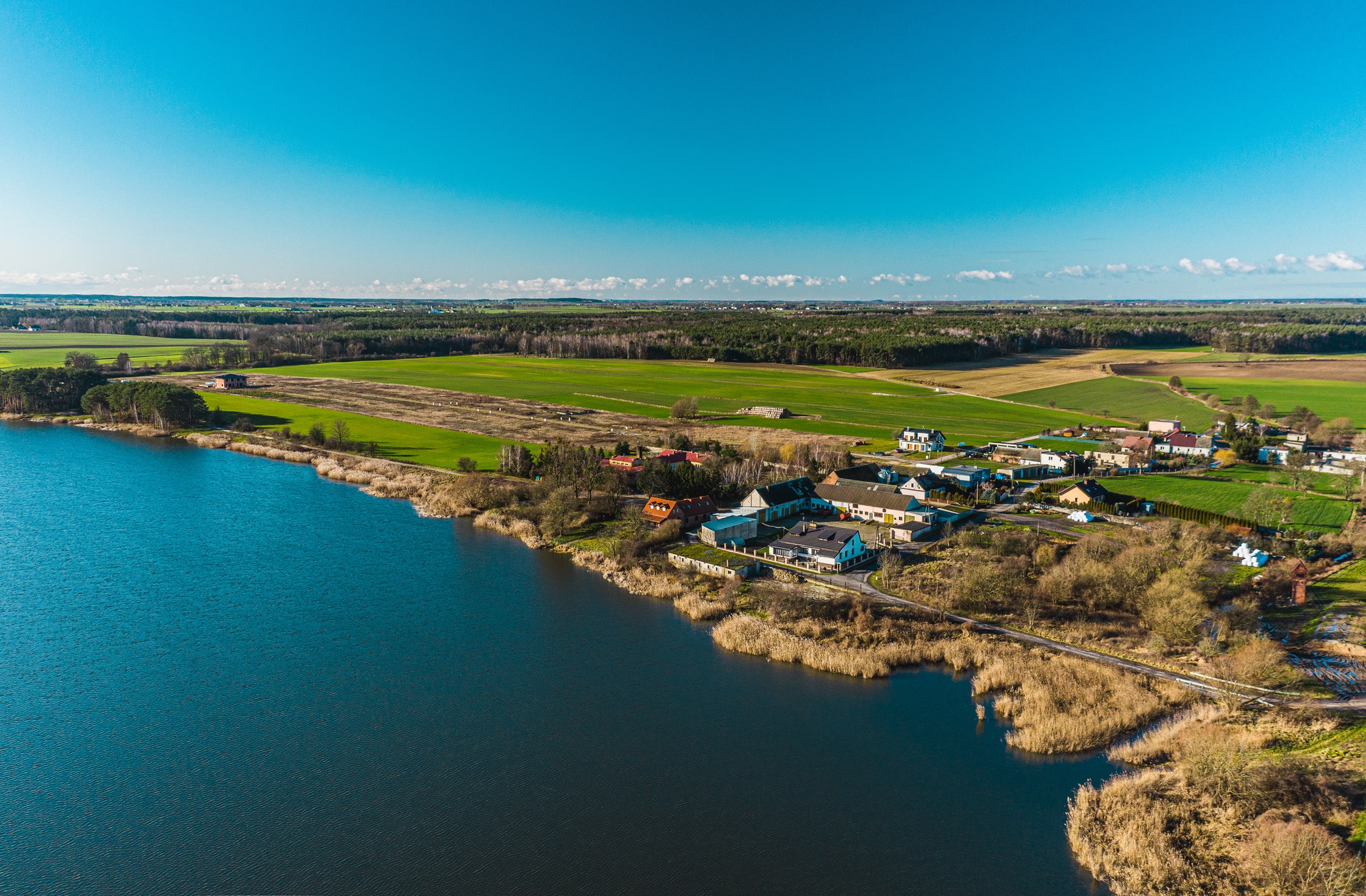 Panoramiczny widok z lotu ptaka na wieś położoną nad jeziorem, z polami uprawnymi w tle, w słoneczny dzień.