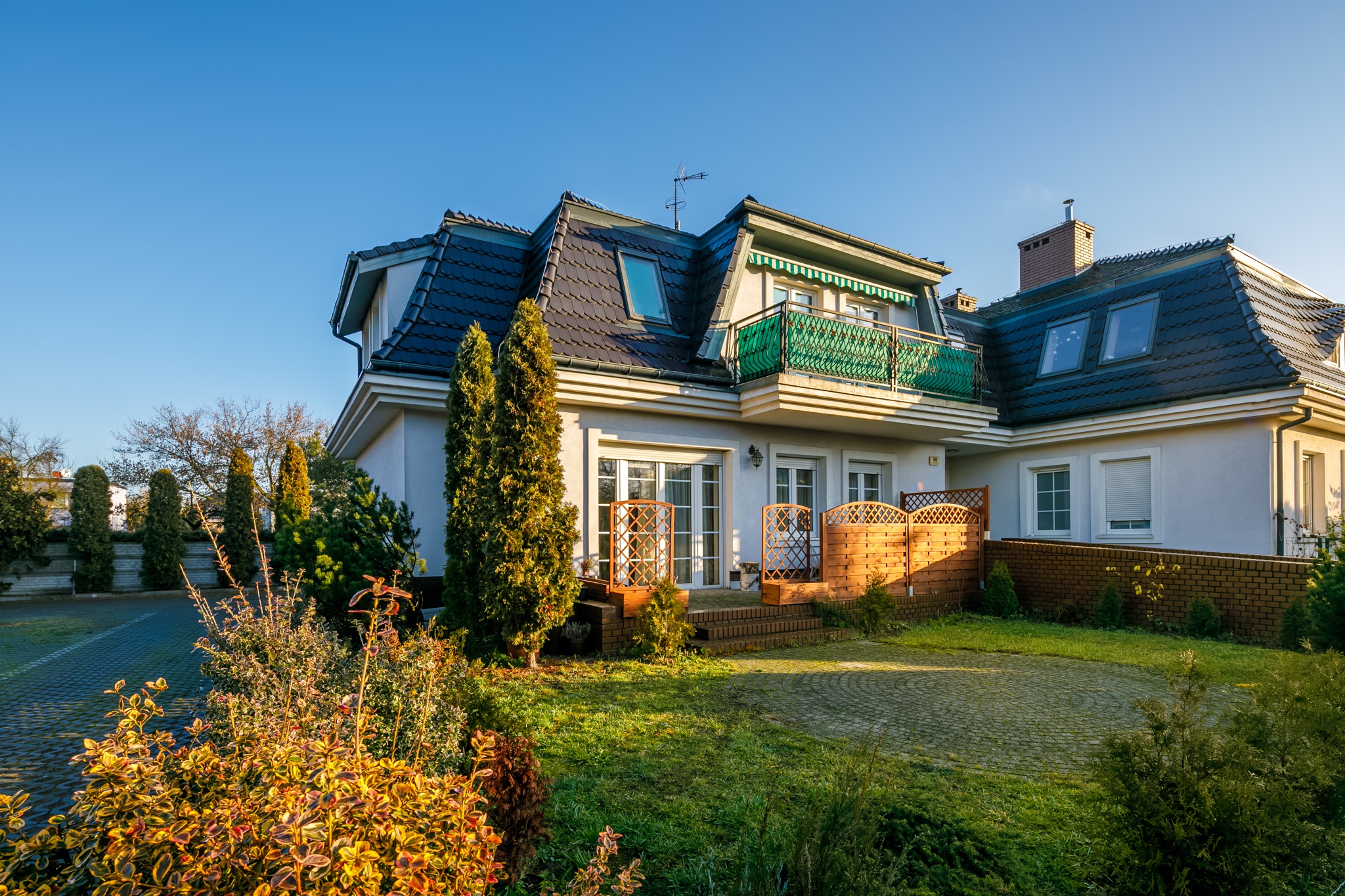 Dwupiętrowy dom z szarego kamienia z ciemnym dachem, balkonem z zieloną balustradą i zadbanym ogrodem z drzewami iglastymi i krzewami.