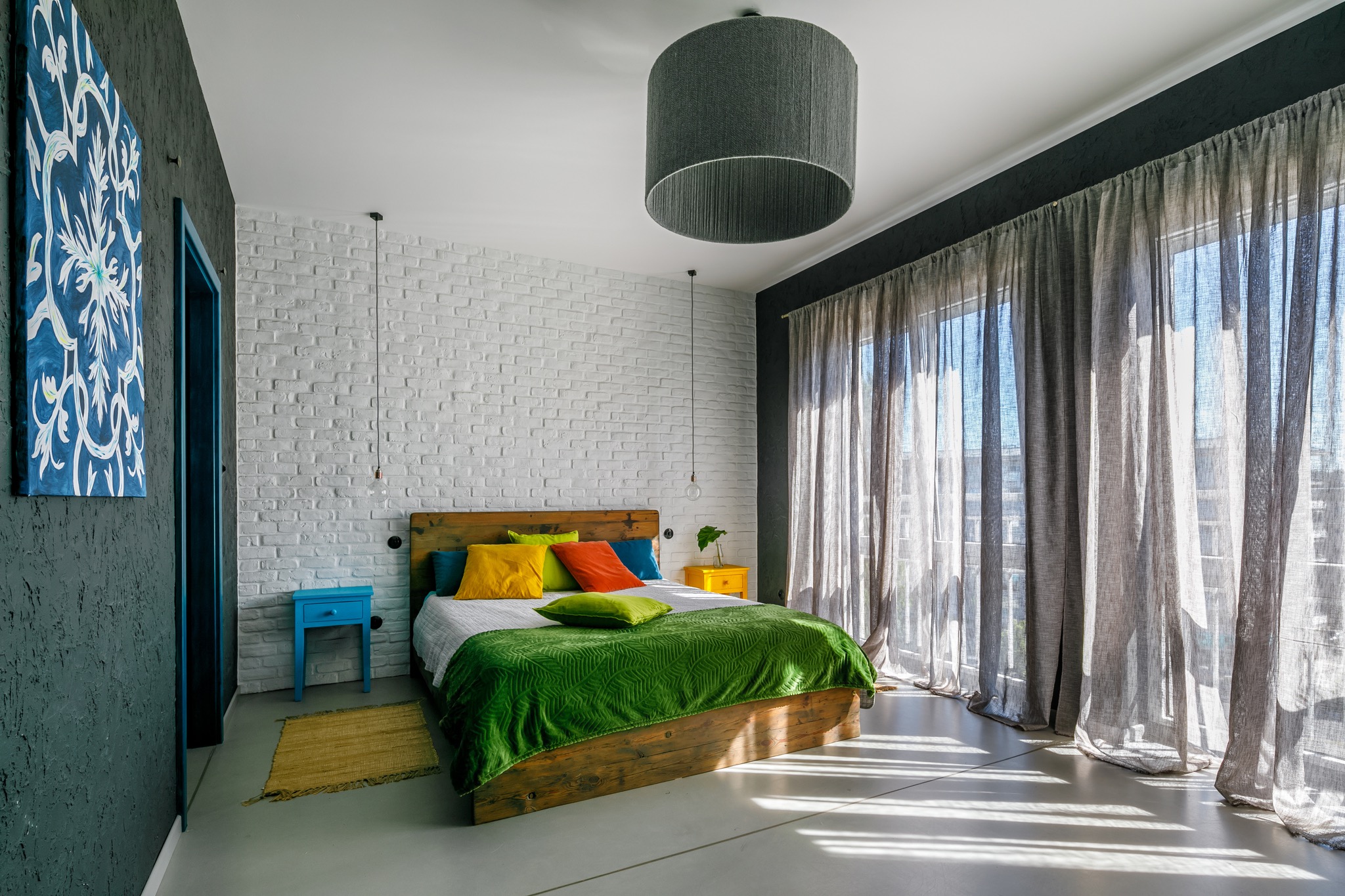 Jasna sypialnia z drewnianym łóżkiem, kolorowymi poduszkami i zielonym pledem, na ścianie biała cegła i abstrakcyjny obraz, obok okna z szarymi zasłonami, na podłodze szary dywanik.