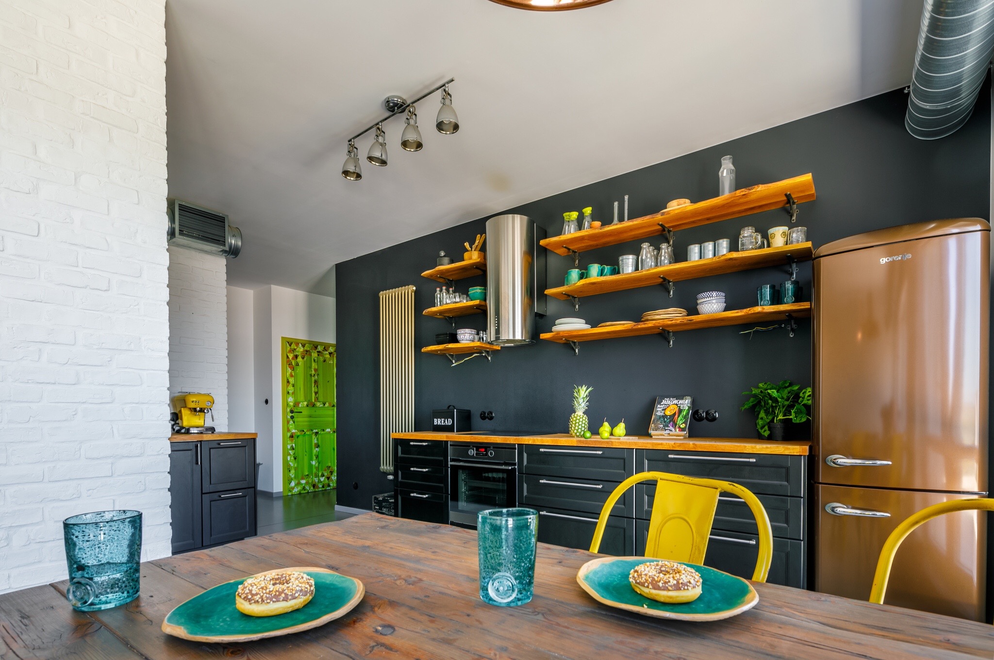 Nowoczesna kuchnia z ciemnymi szafkami, drewnianymi półkami z dekoracjami, miedzianą lodówką Gorenje i drewnianym stołem z dwoma pączkami na turkusowych talerzach oraz szklankami w kolorze morza.