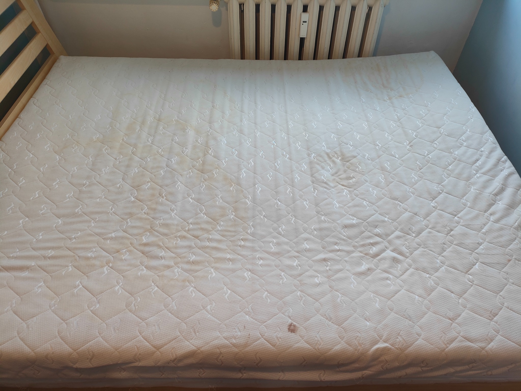 Biały materac z widocznymi plamami i zabrudzeniami, leżący na drewnianej ramie łóżka, z grzejnikiem w tle.