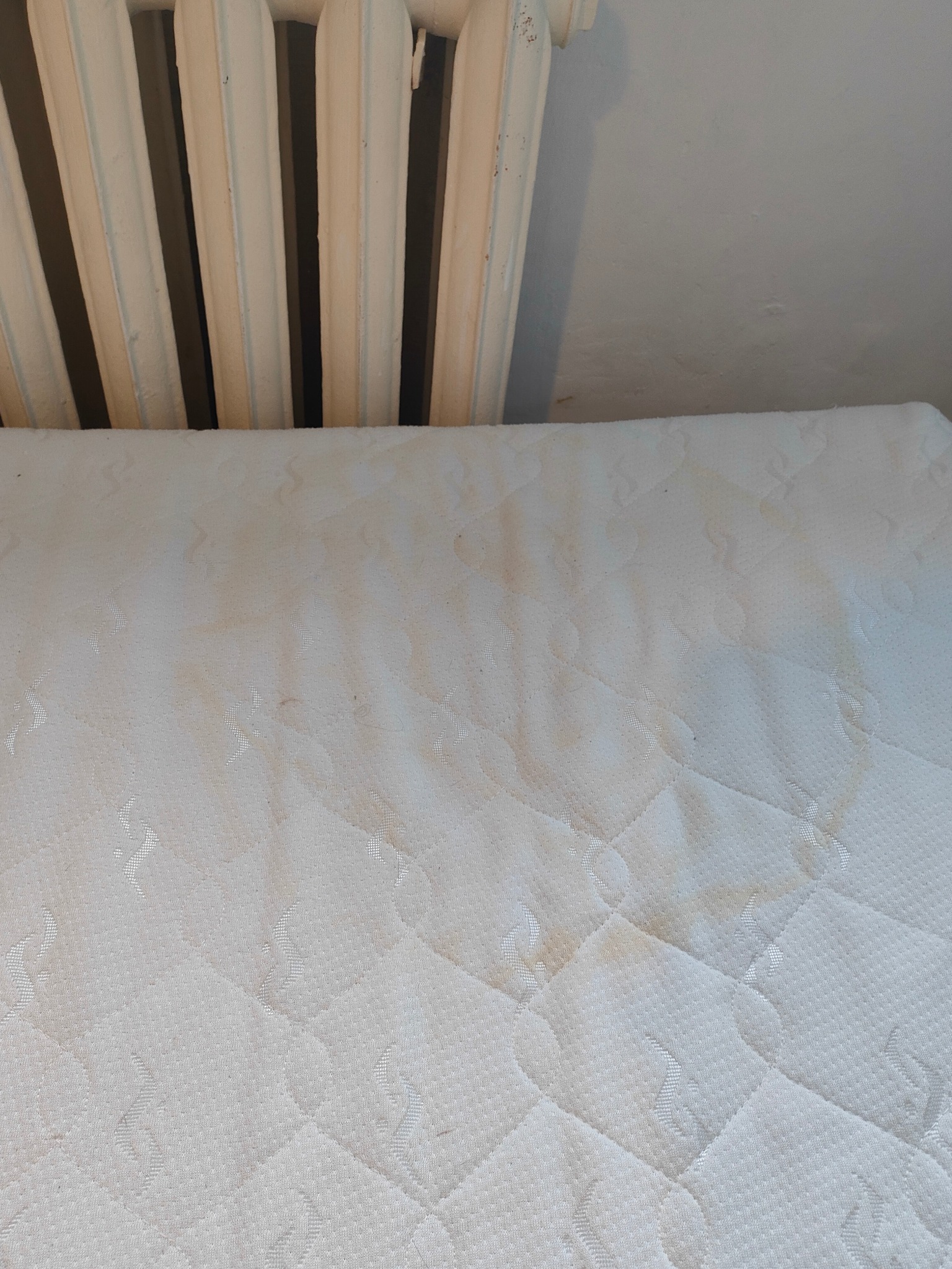 Mocno zabrudzony materac w pobliżu starego, żeliwnego grzejnika.