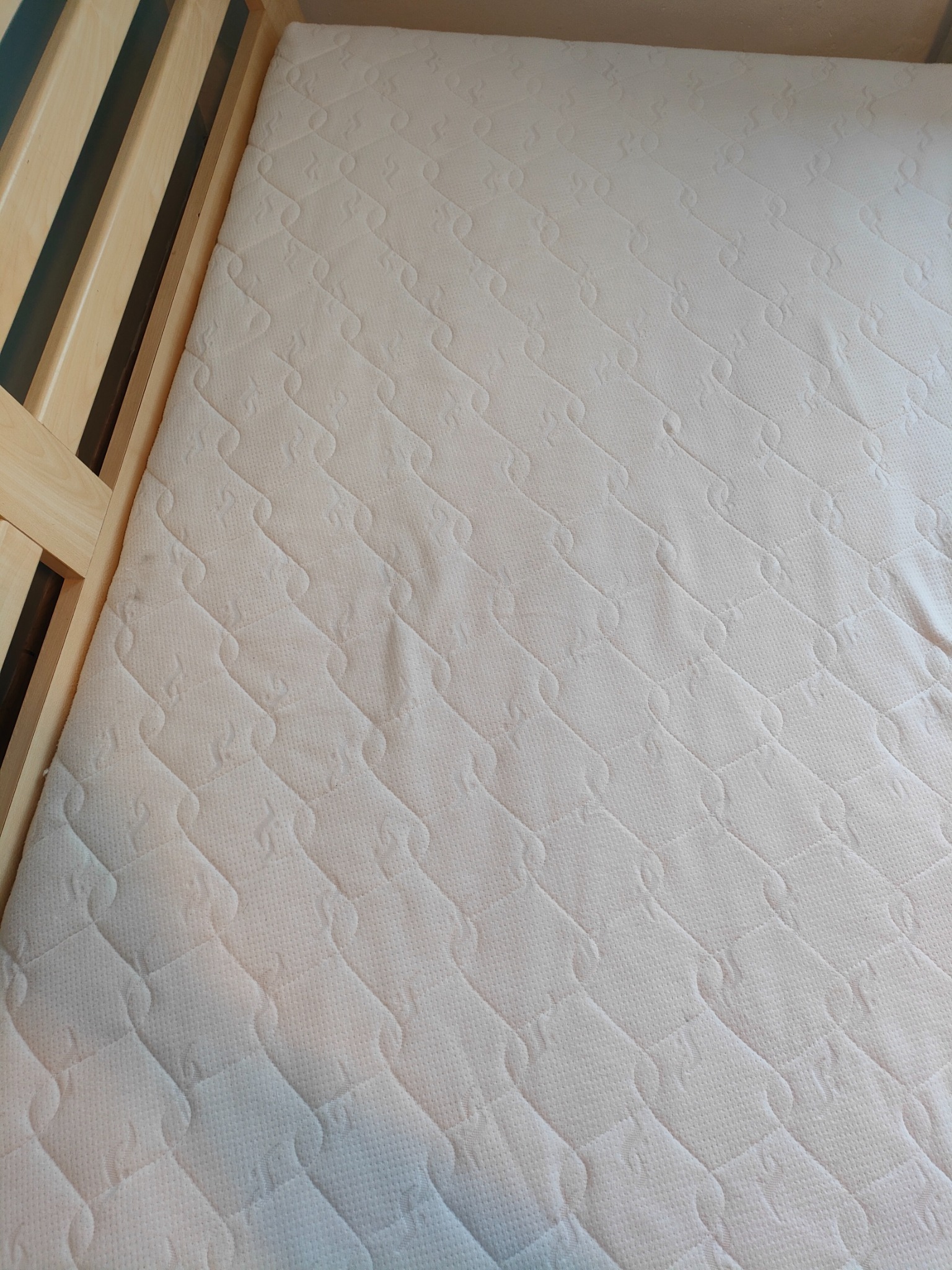 Jasny materac z pikowanym wzorem w drewnianej ramie łóżka, widoczne delikatne cieniowanie na powierzchni.