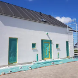 Biały dom w trakcie ocieplania, z widocznym rusztowaniem, zabezpieczonymi folią oknami i drzwiami, oraz fragmentem ocieplonej ściany z widoczną warstwą izolacyjną.