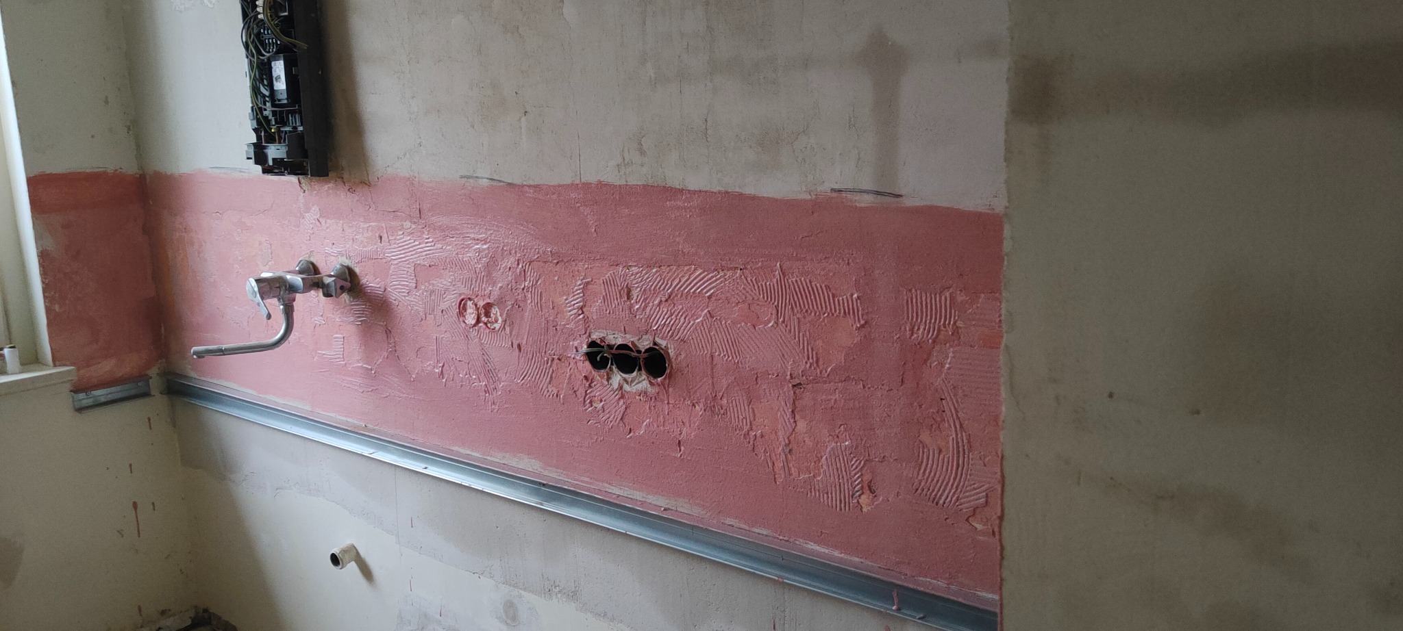 Przygotowana ściana pod płytki w łazience z widocznymi otworami na gniazdka elektryczne i rurę wodociągową, nałożony różowy podkład pod płytki oraz metalowy profil przy podłodze.