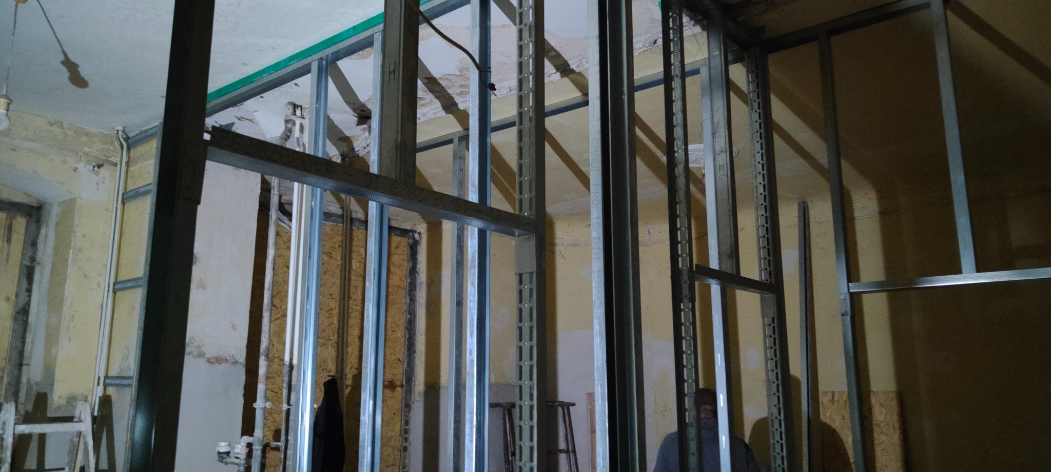 Stelaż z profili metalowych w trakcie montażu w pomieszczeniu z widoczną starą instalacją elektryczną i resztkami tapety, przygotowanie do dalszych prac wykończeniowych.