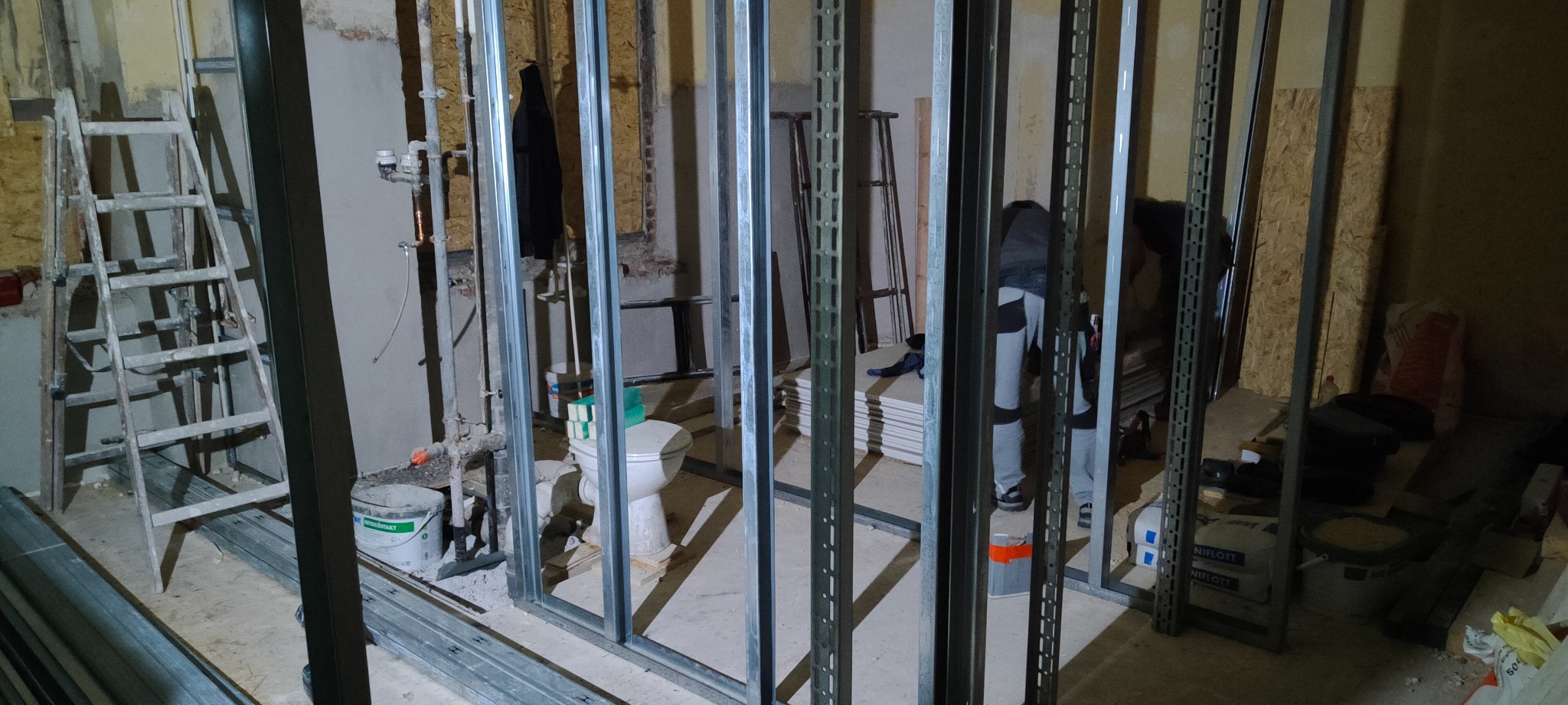 Montaż stelaża z profili metalowych w łazience, widoczna toaleta, drabina i materiały budowlane w surowym stanie.