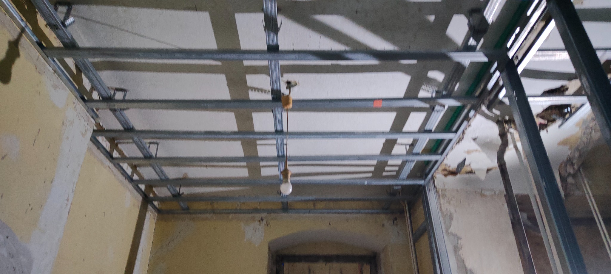 Metalowa konstrukcja sufitu podwieszanego w trakcie montażu, widoczne profile i wisząca żarówka, ściany w stanie surowym z widocznymi śladami po starych powłokach.