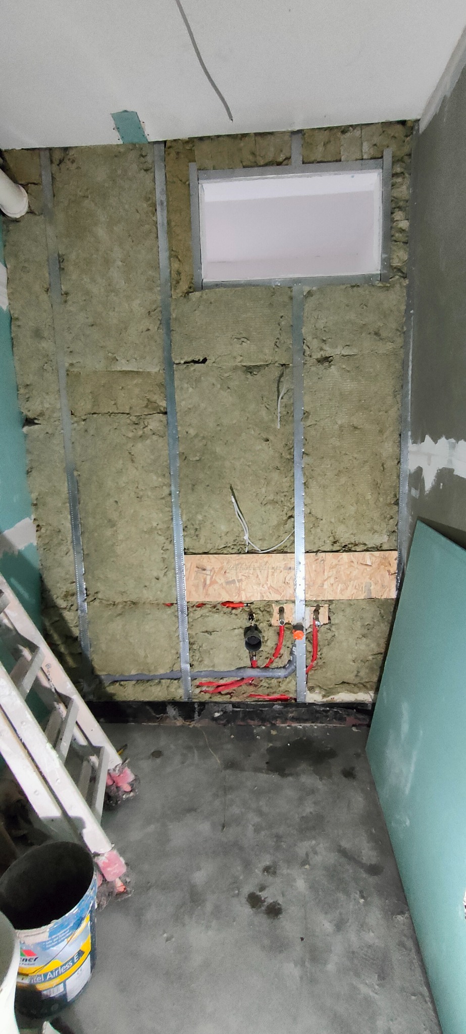 Łazienka w trakcie remontu: ocieplona ściana z wełny mineralnej, metalowy stelaż, fragment instalacji wodnej z czerwonymi rurkami, drabina i wiadro z farbą w tle.