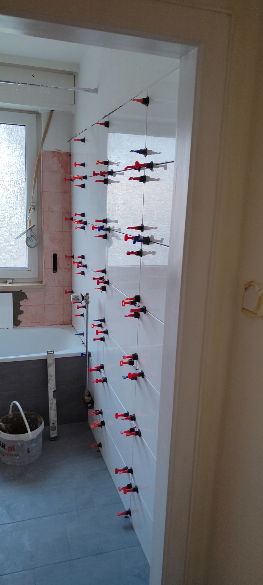 Proces układania białych płytek ściennych w łazience z użyciem systemów poziomowania, widoczny fragment wanny i kubeł z zaprawą.